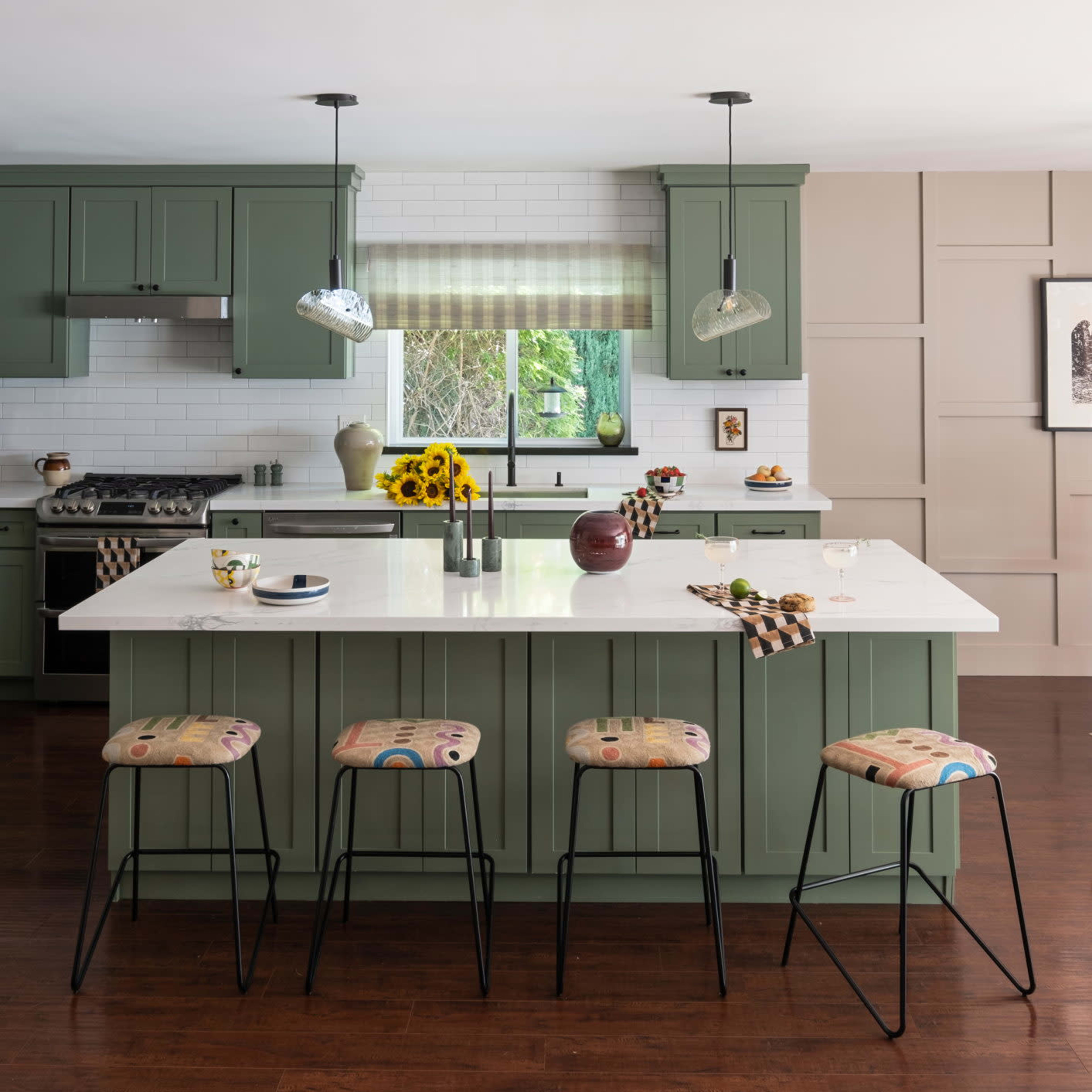 The kitchen features green cabinetry, a large white island with bar stools, and a window overlooking a garden filled with sunflowers.