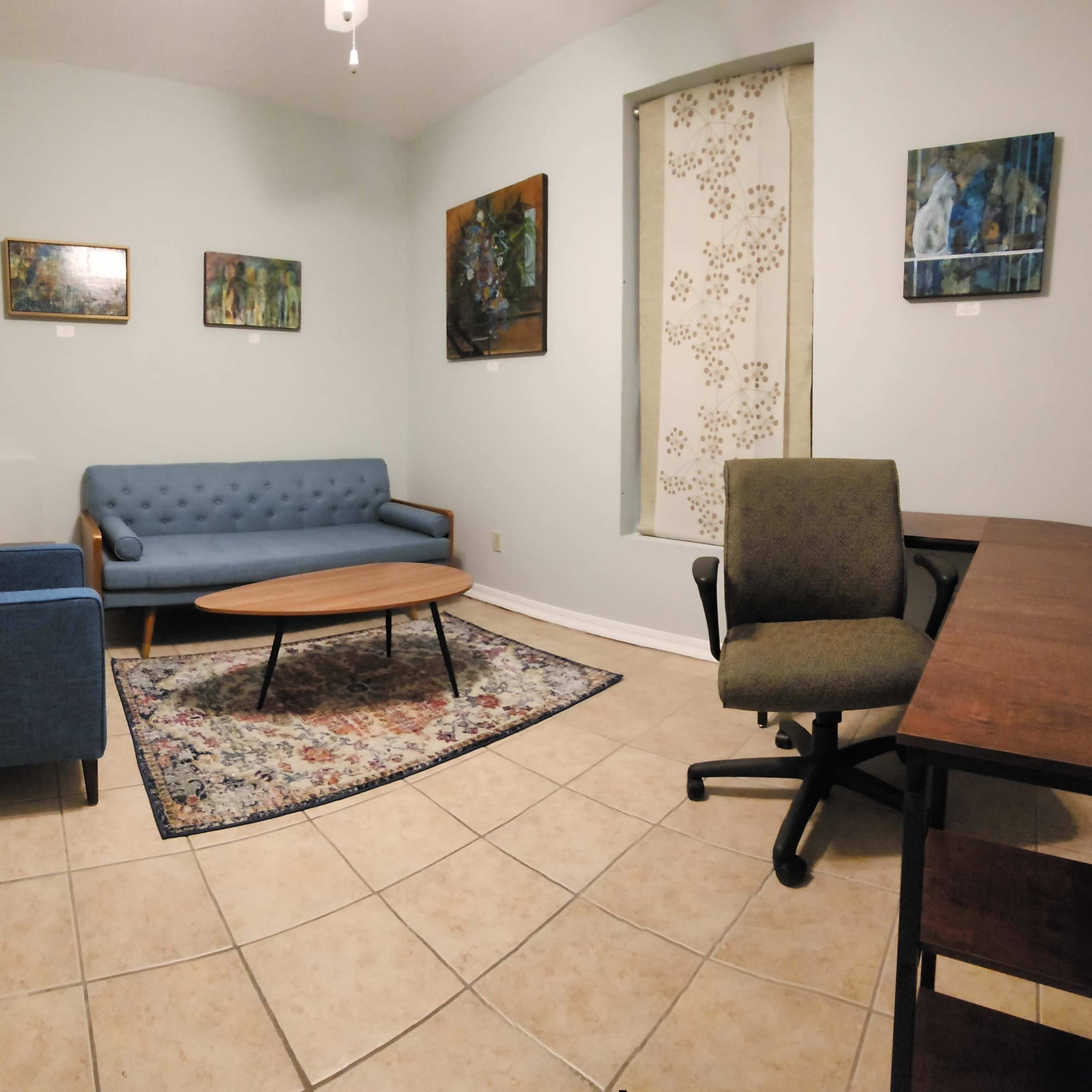 A small, well-lit waiting room with two blue couches, a round coffee table, a patterned rug, a desk with a chair, and several framed paintings on the walls.