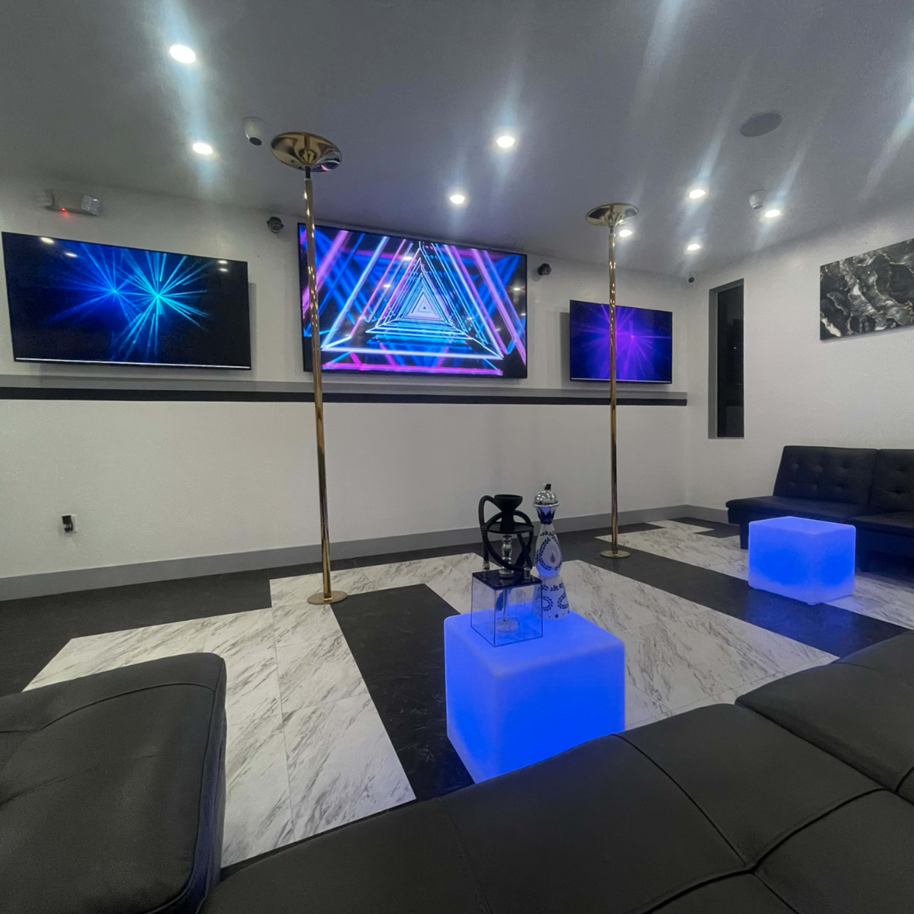 A modern lounge area featuring two television screens, sleek black couches, and illuminated blue tables.