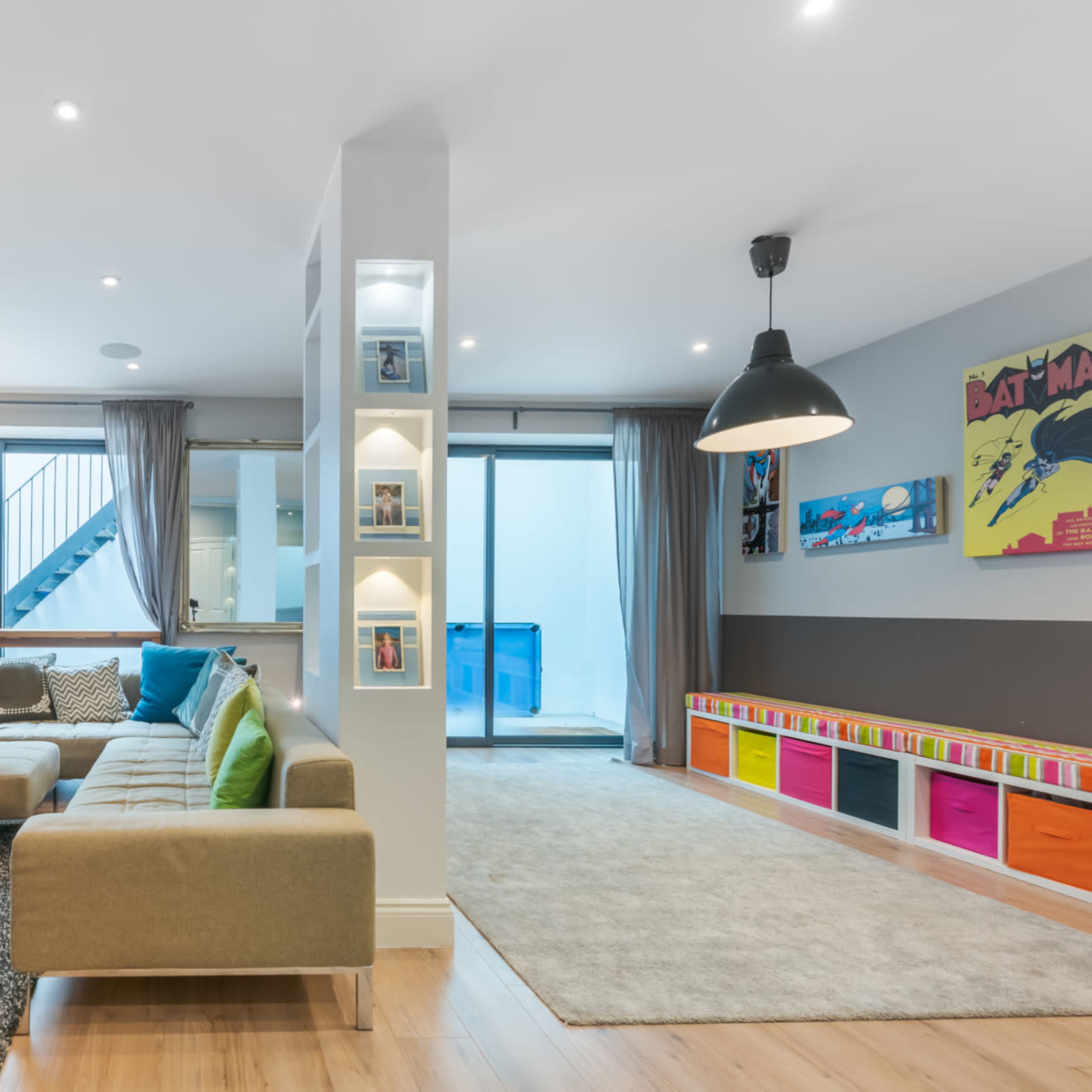 A modern living room with a sectional sofa, a television unit, and colorful storage bins along one wall, adjacent to a staircase and a large window.