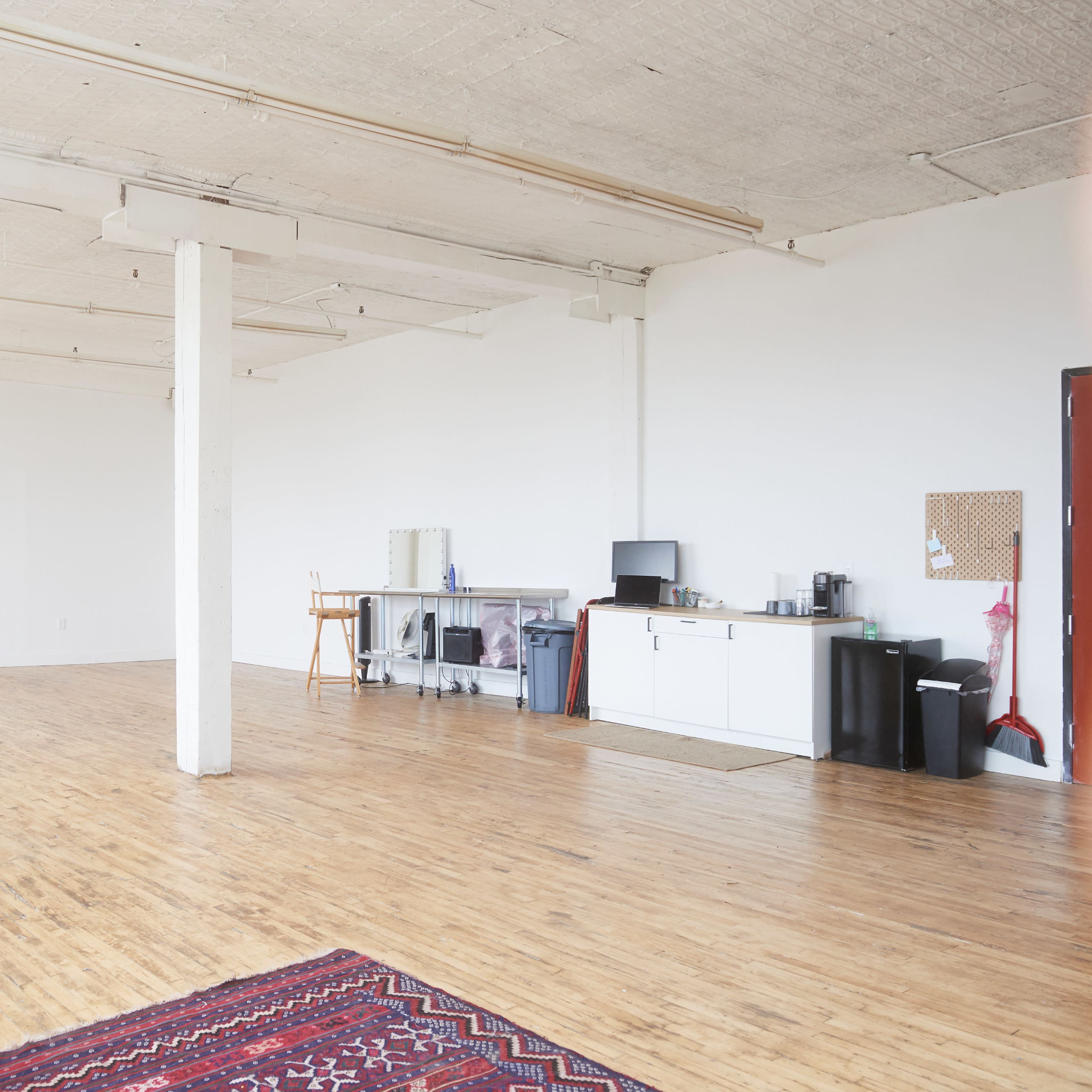 A spacious, empty room with wooden flooring, white walls, and a small kitchenette in the corner.