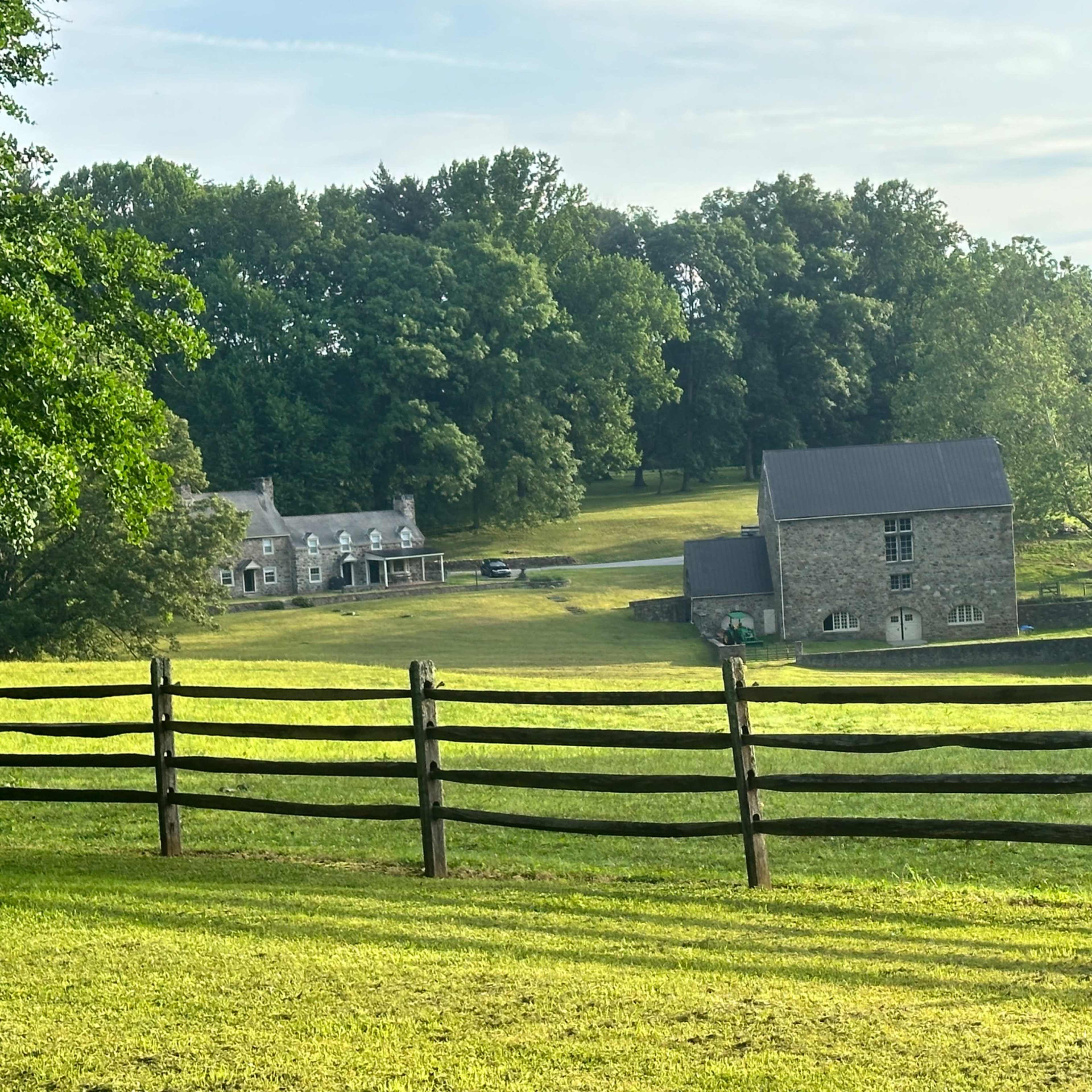 A stone house and a separate building set against a backdrop of lush green trees and a wooden fence.
