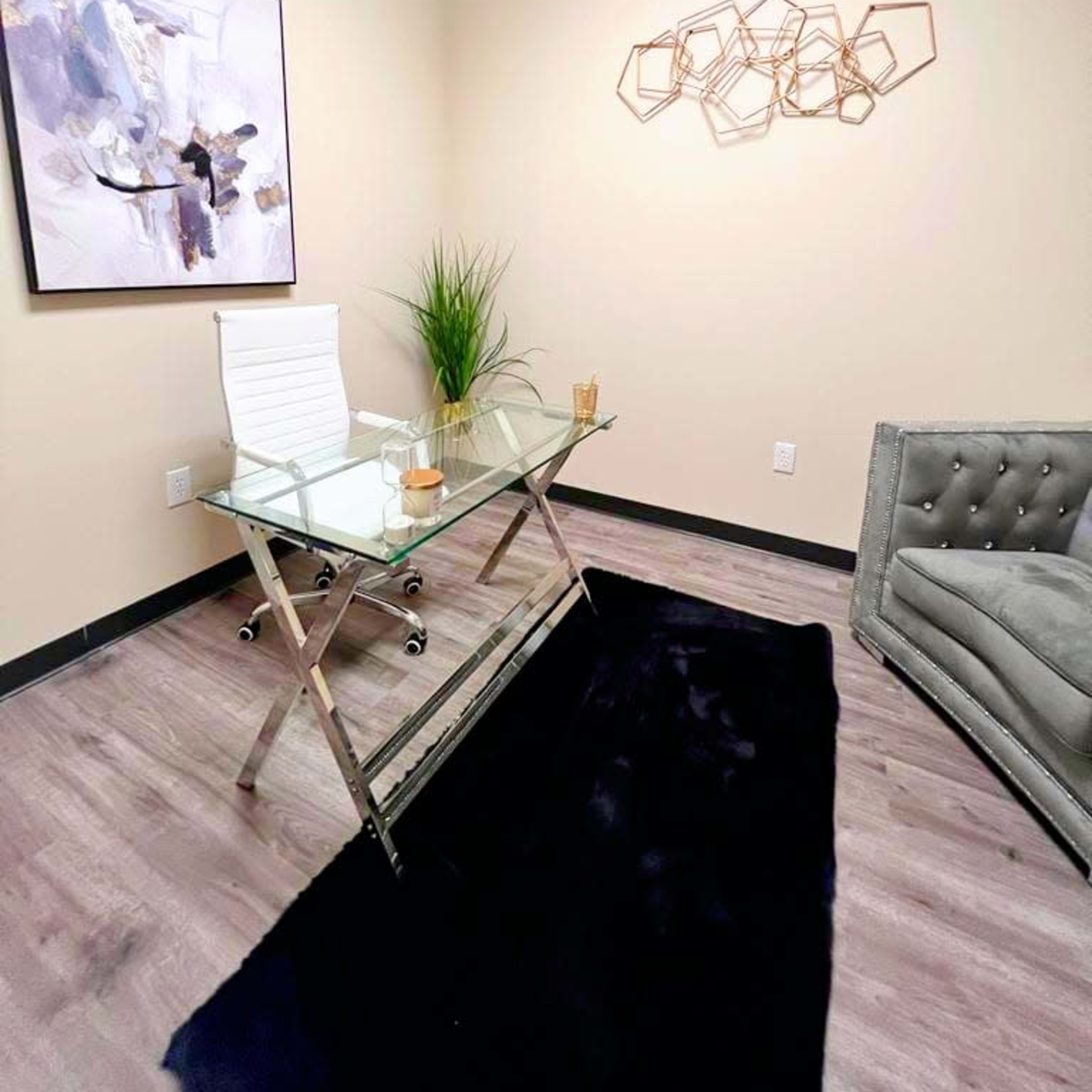 A modern office space with a glass desk, a white chair, a gray tufted sofa, a black rug, and decorative wall art.