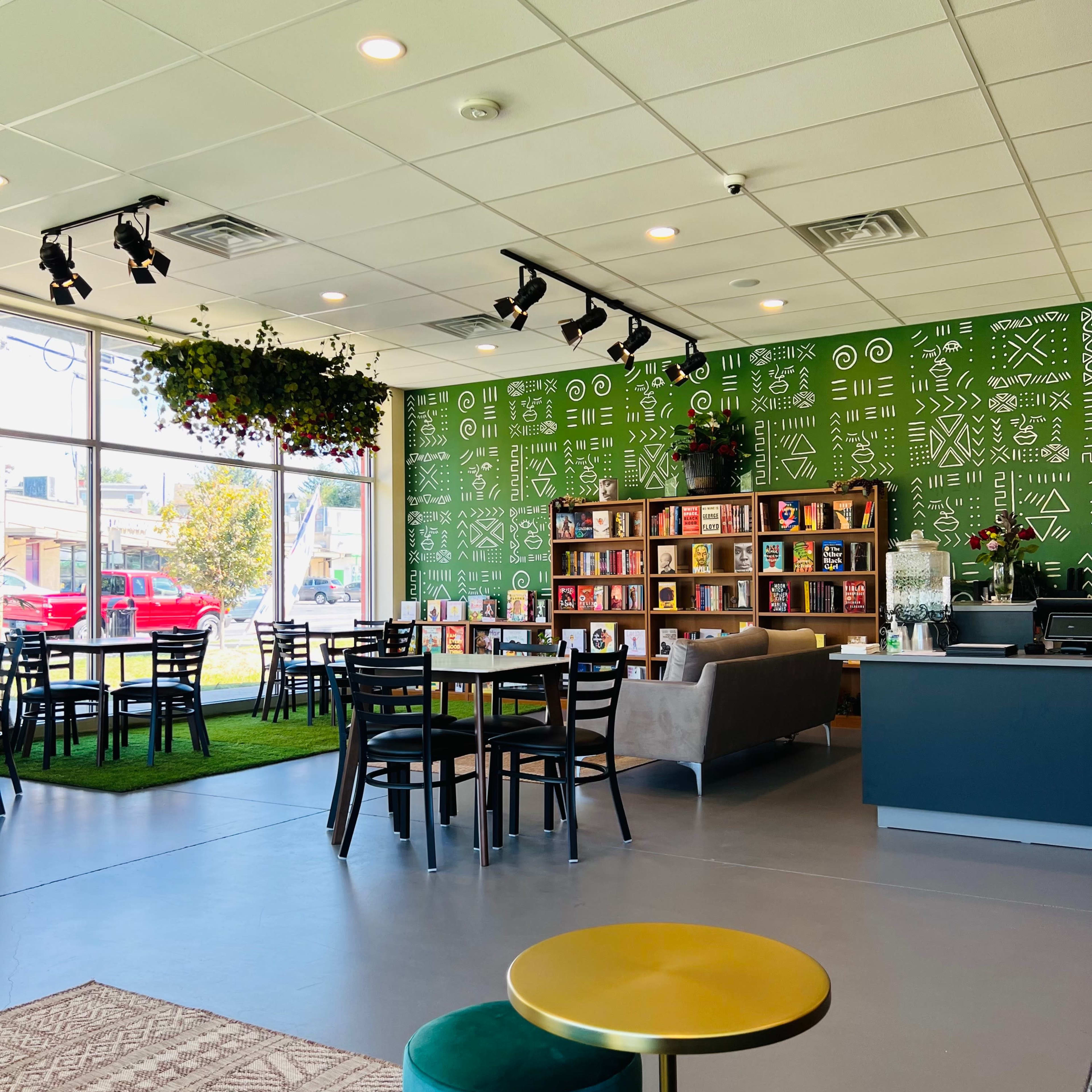 A modern cafe interior with green patterned walls, wooden bookshelves filled with books, tables and chairs, and a cozy seating area.