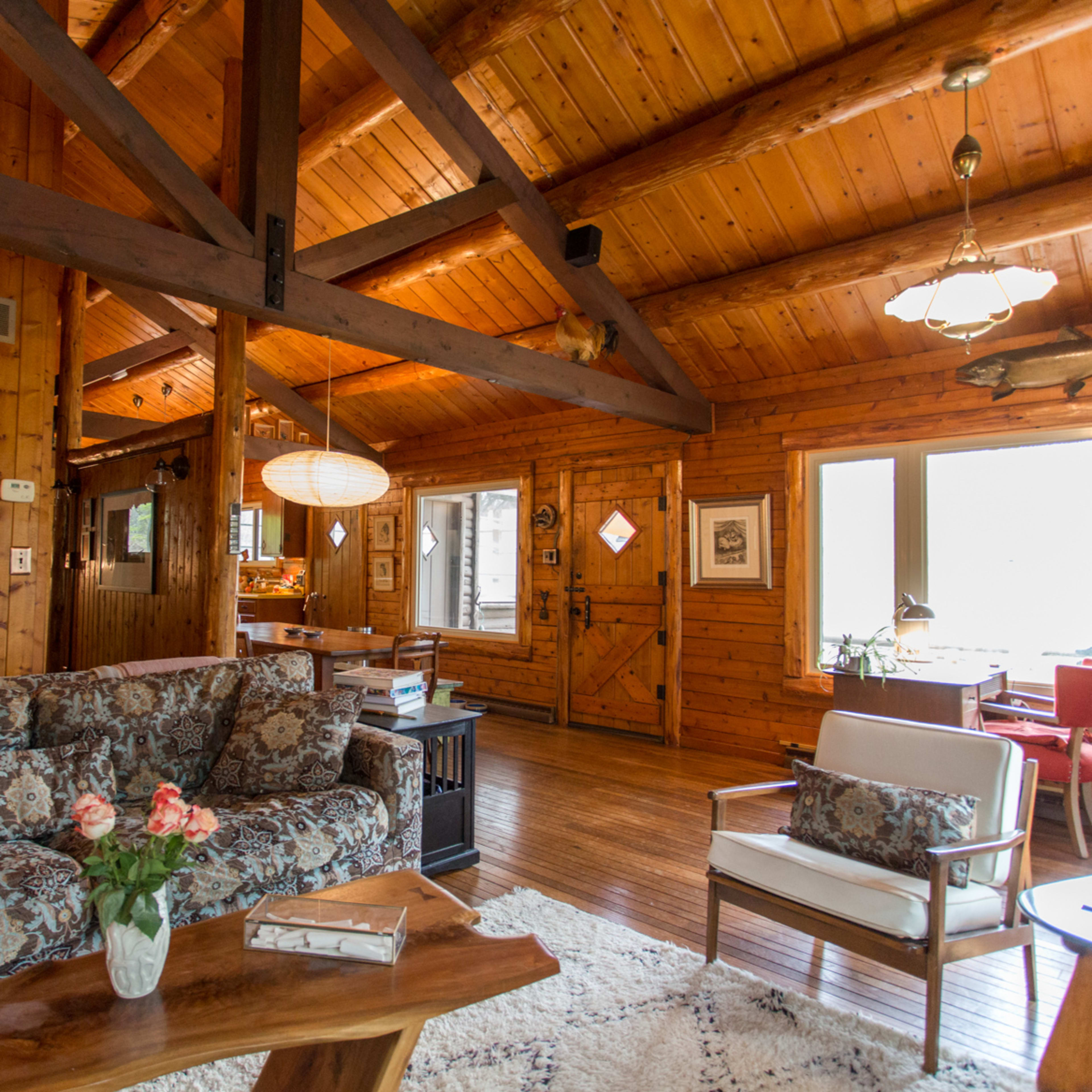 A wooden cabin interior with a combination of a living area featuring a patterned sofa, a coffee table, and large windows allowing natural light.