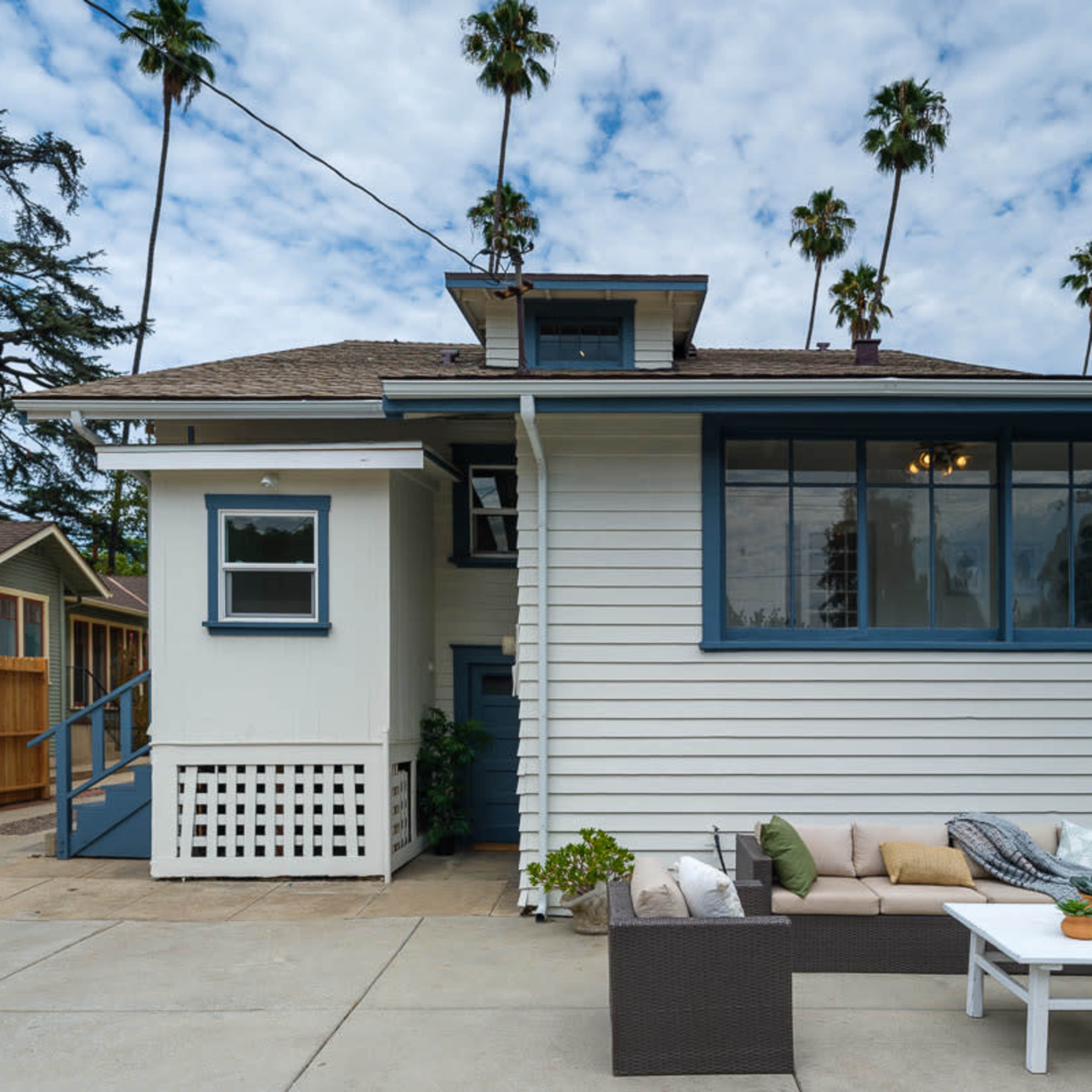 A two-story house with a screened porch and a patio area furnished with a sofa and coffee table, surrounded by palm trees.