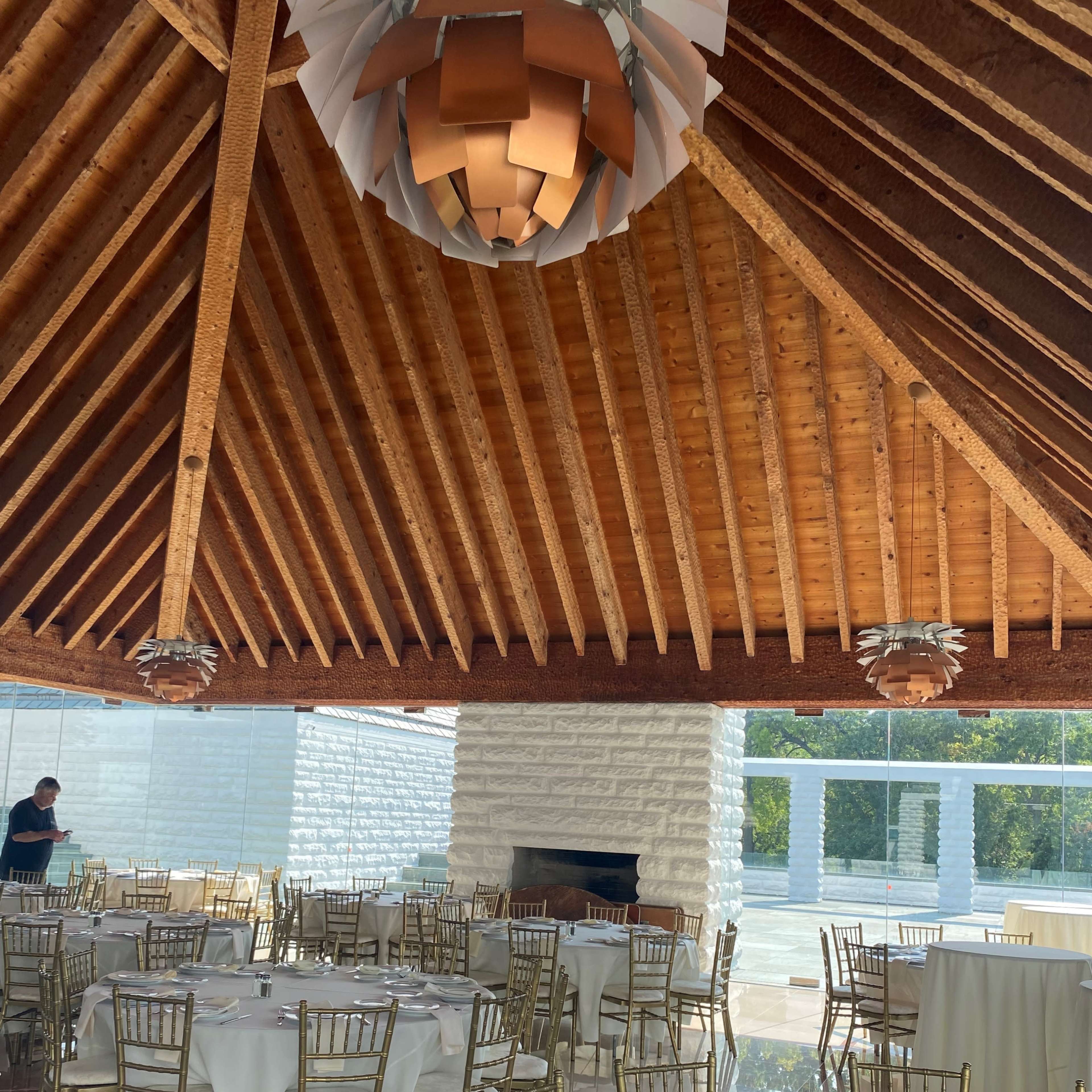 A spacious dining area with round tables set for a meal, featuring wooden beams and modern light fixtures overhead.