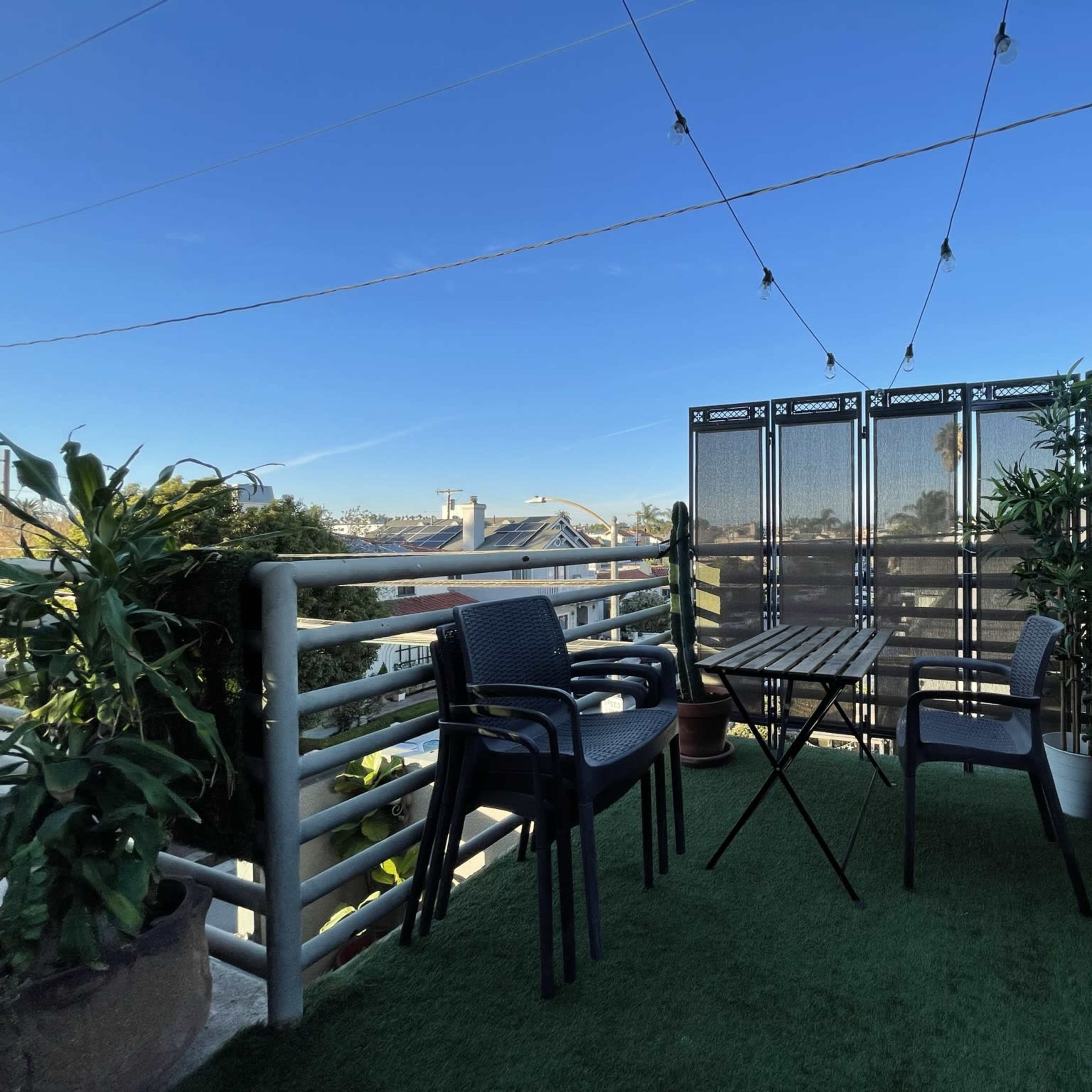 A balcony with two black chairs, a small table, potted plants, and a decorative screen, all set against a clear blue sky.