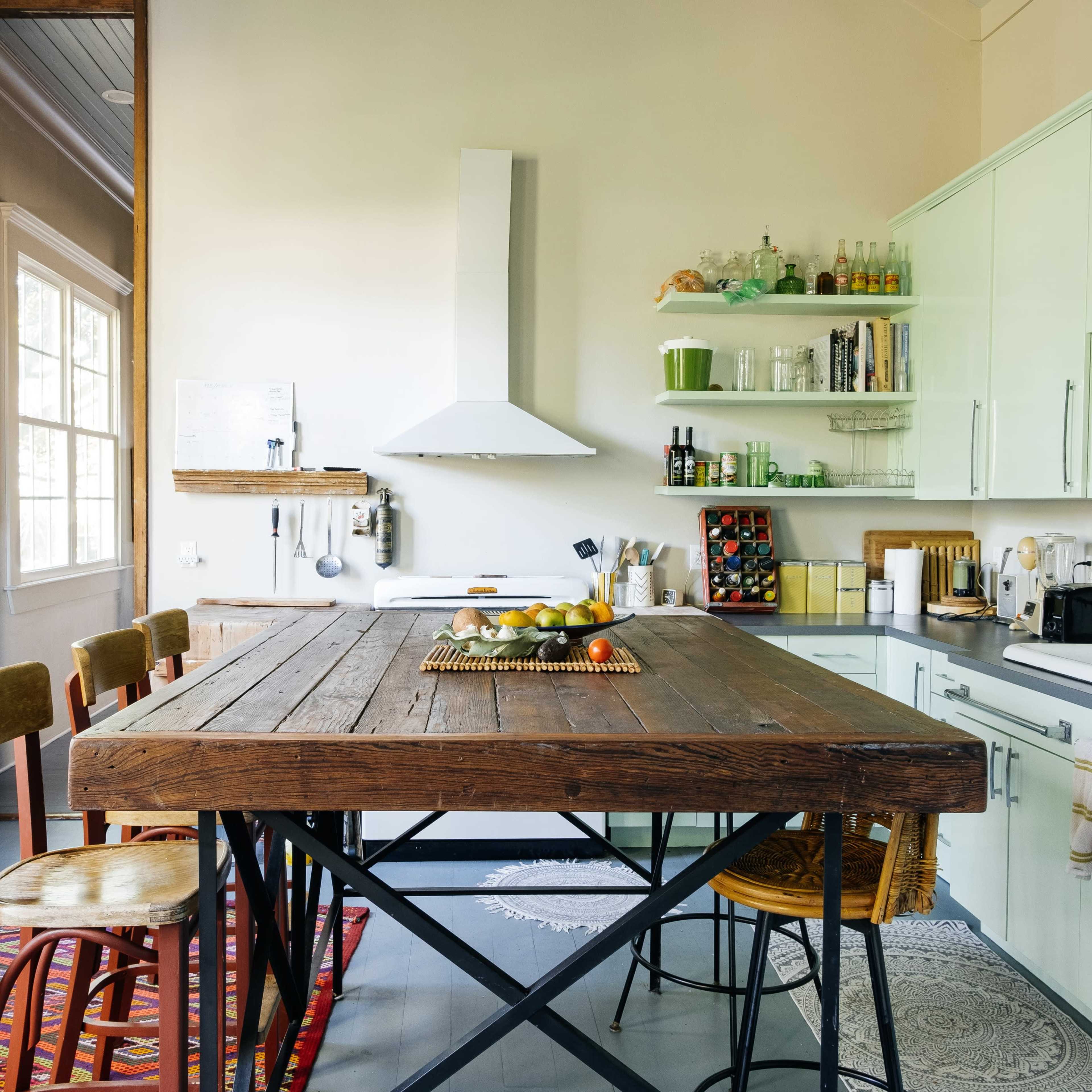 A modern kitchen features a large wooden table, green cabinetry, and an array of kitchen tools and ingredients organized on shelves.