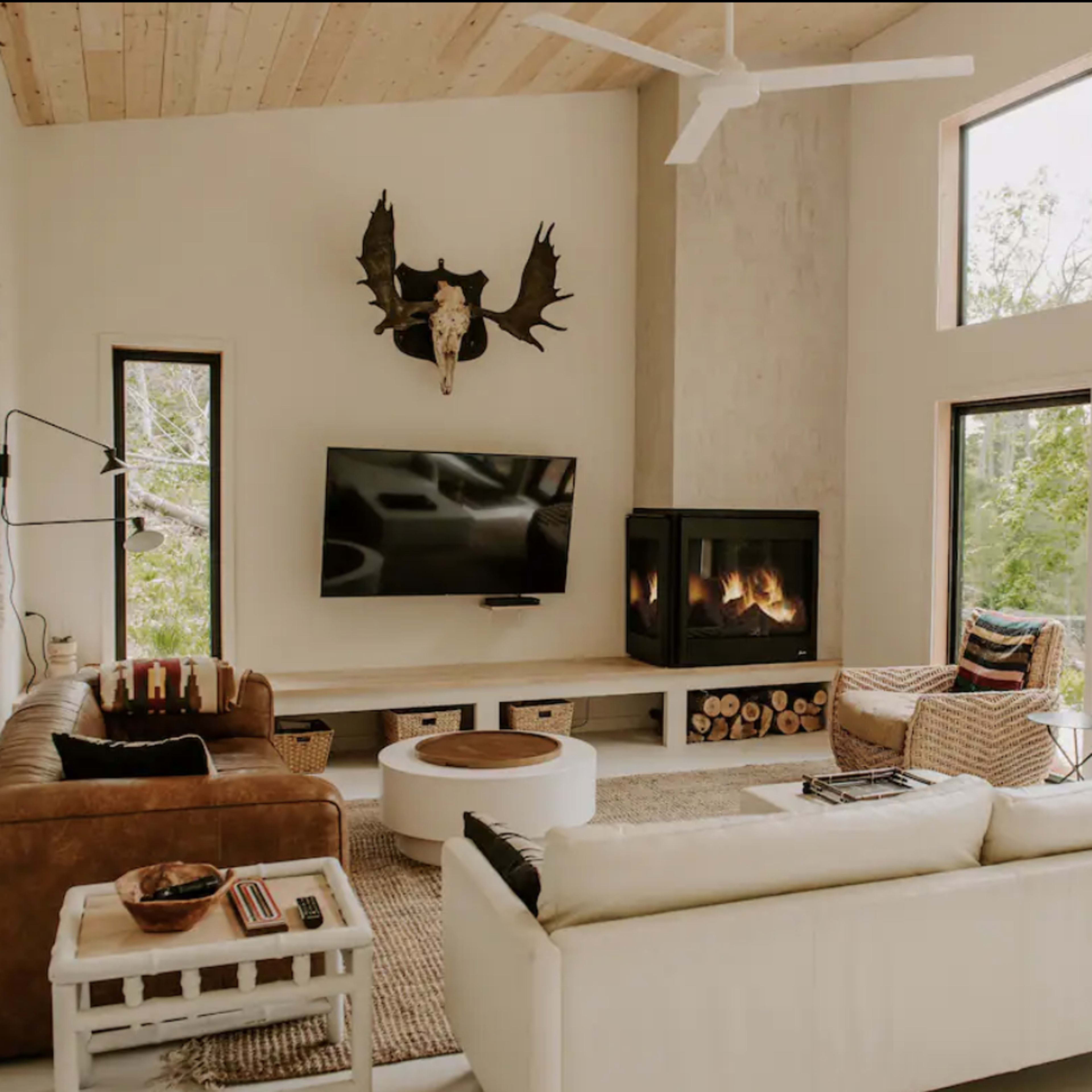 A modern living room features large windows, a mounted moose head, a flat-screen TV, a fireplace, and both leather and white sofas.