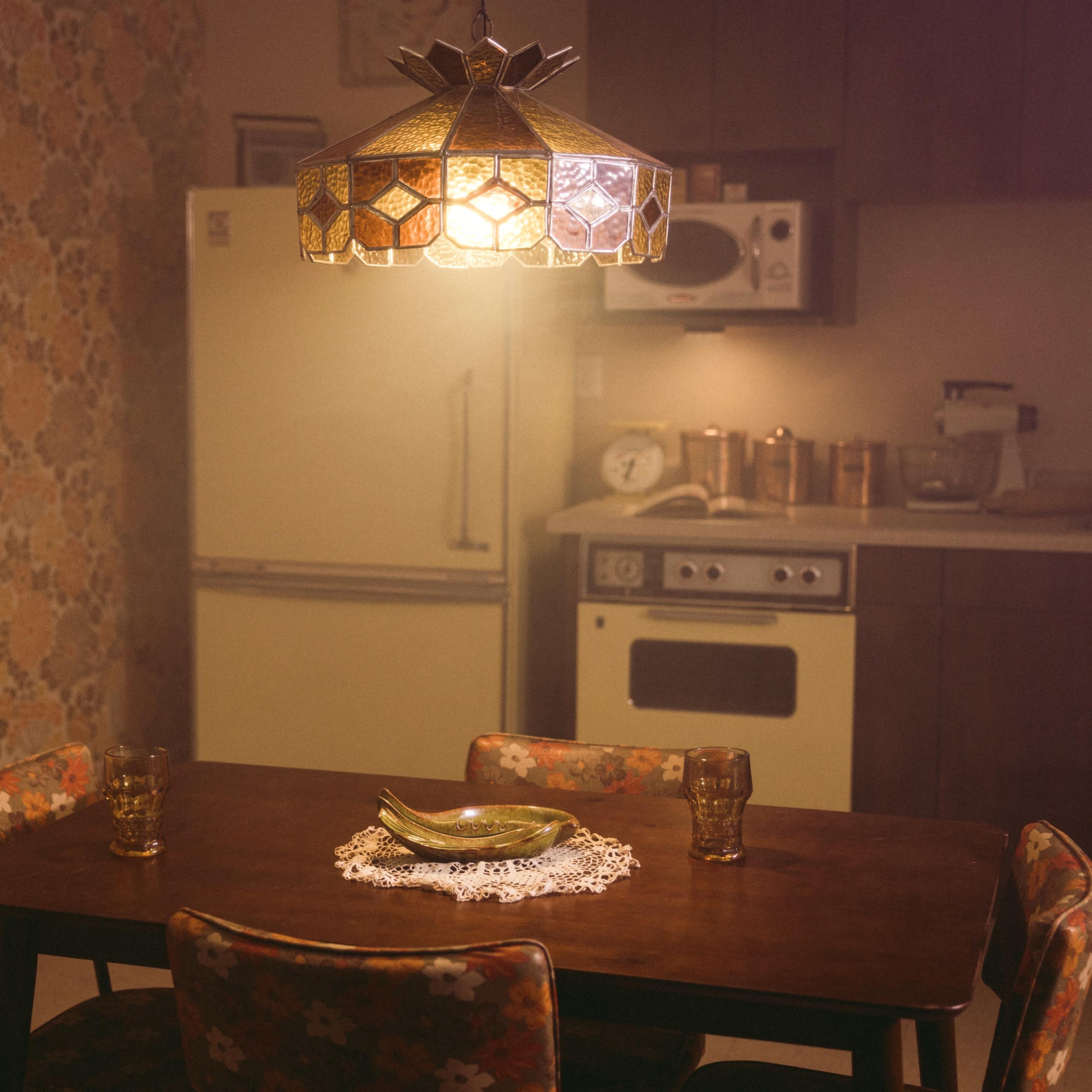 A vintage kitchen features a wooden dining table set with a bowl and glasses under a patterned lampshade, surrounded by retro wallpaper and kitchen appliances.