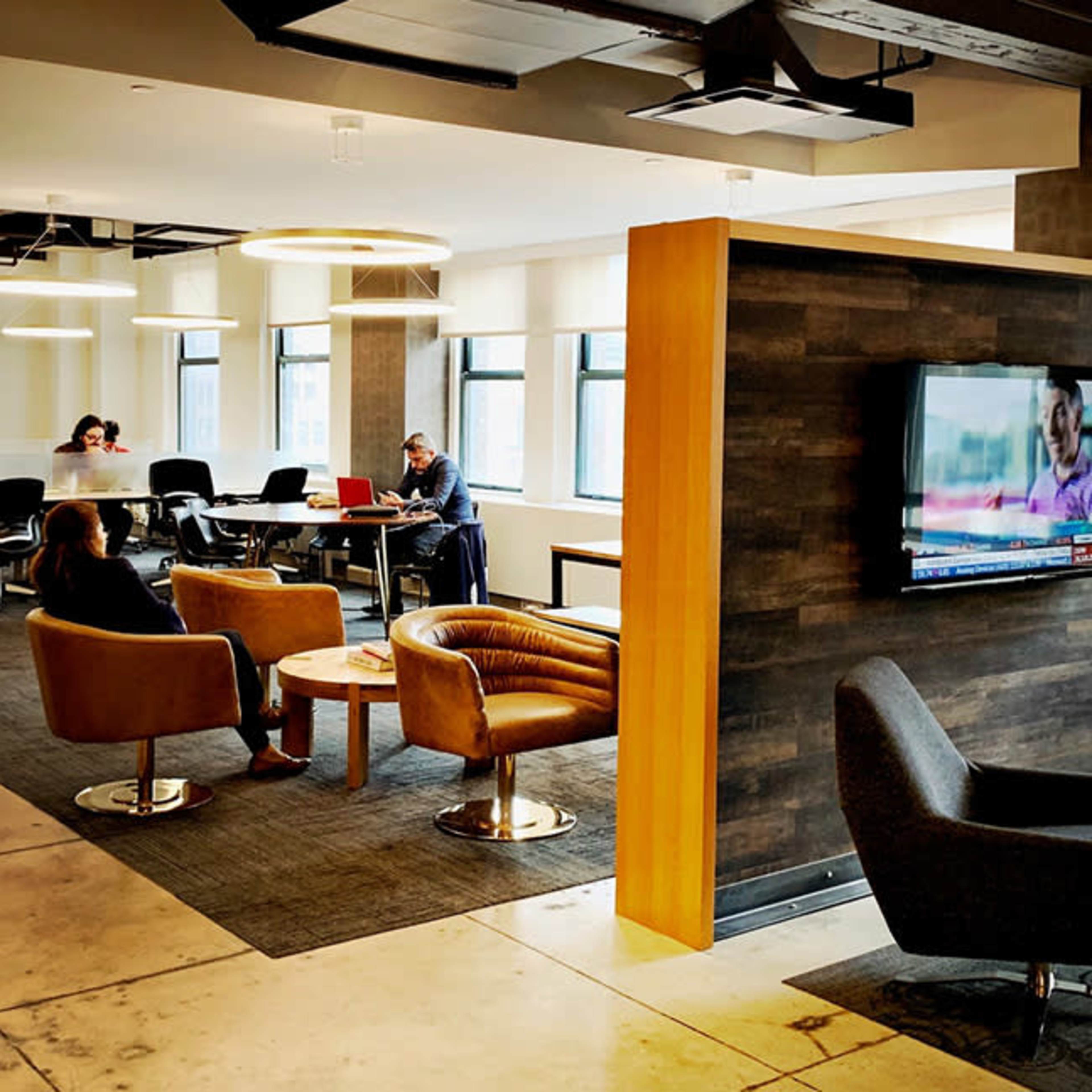 An open office space features various seating arrangements and a large table, with a television mounted on a wooden partition.