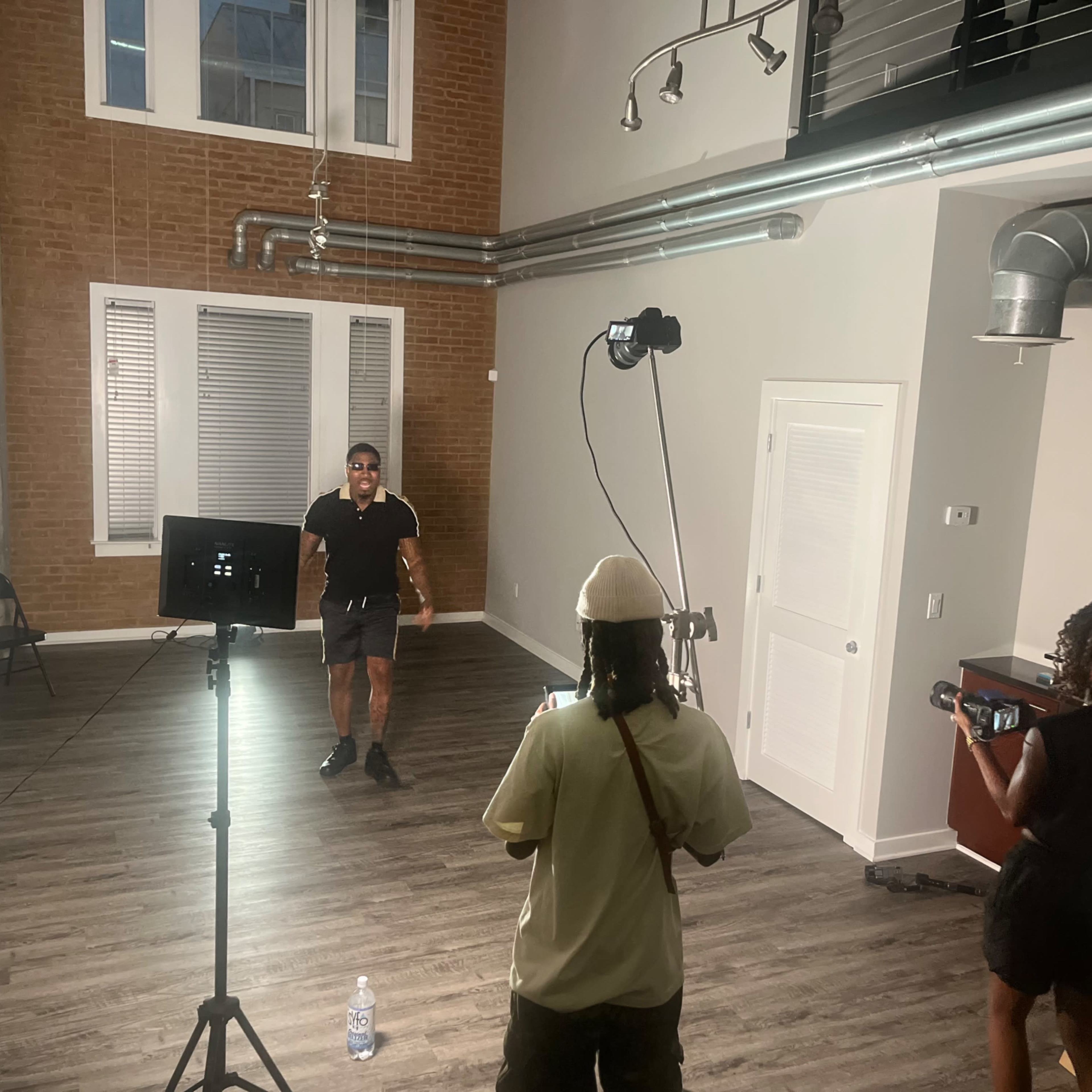 A man stands in the center of an empty, modern room with tall windows, while a woman captures footage with a camera and another person adjusts a light.