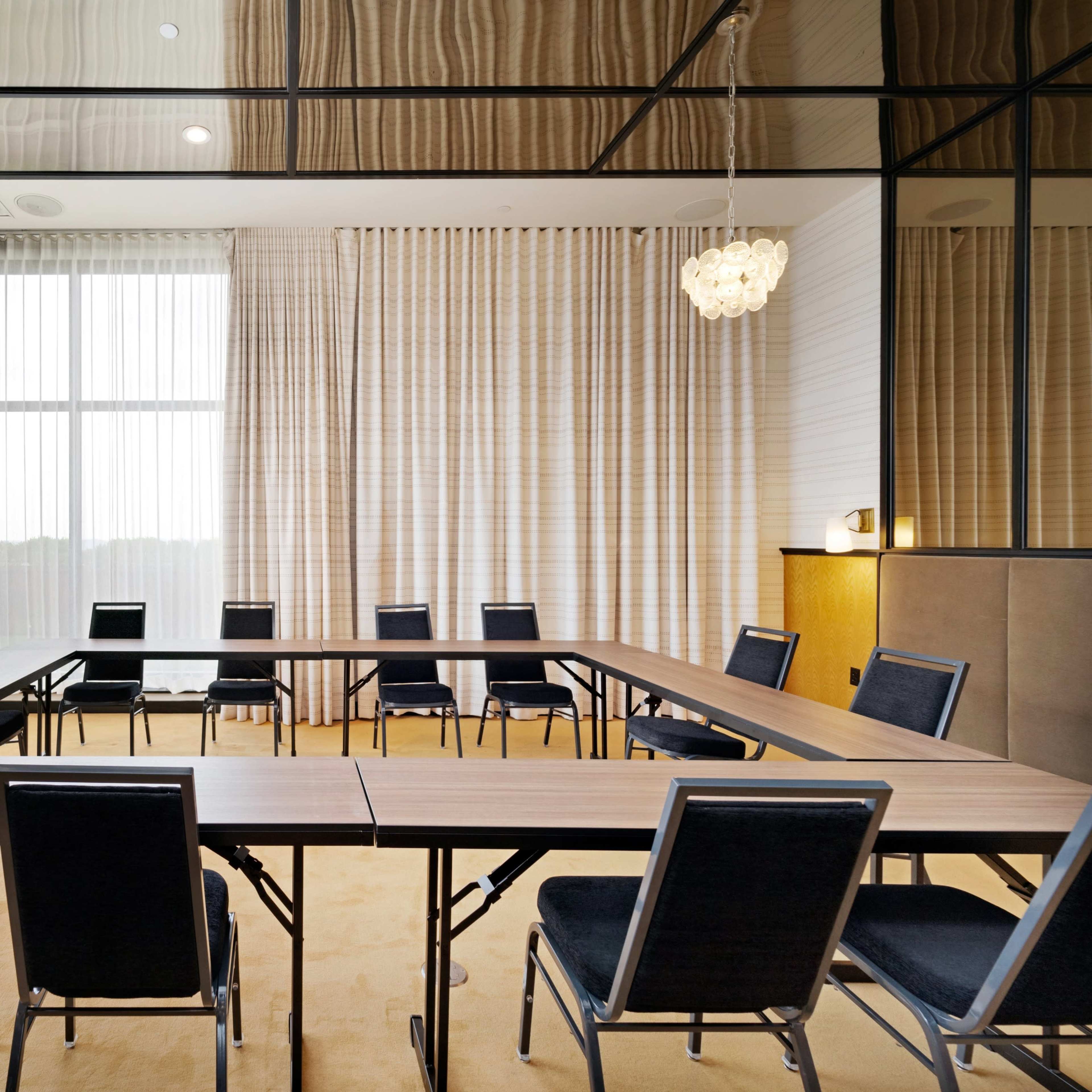 A conference room features a rectangular arrangement of wooden tables surrounded by black chairs, with large windows allowing natural light to enter.
