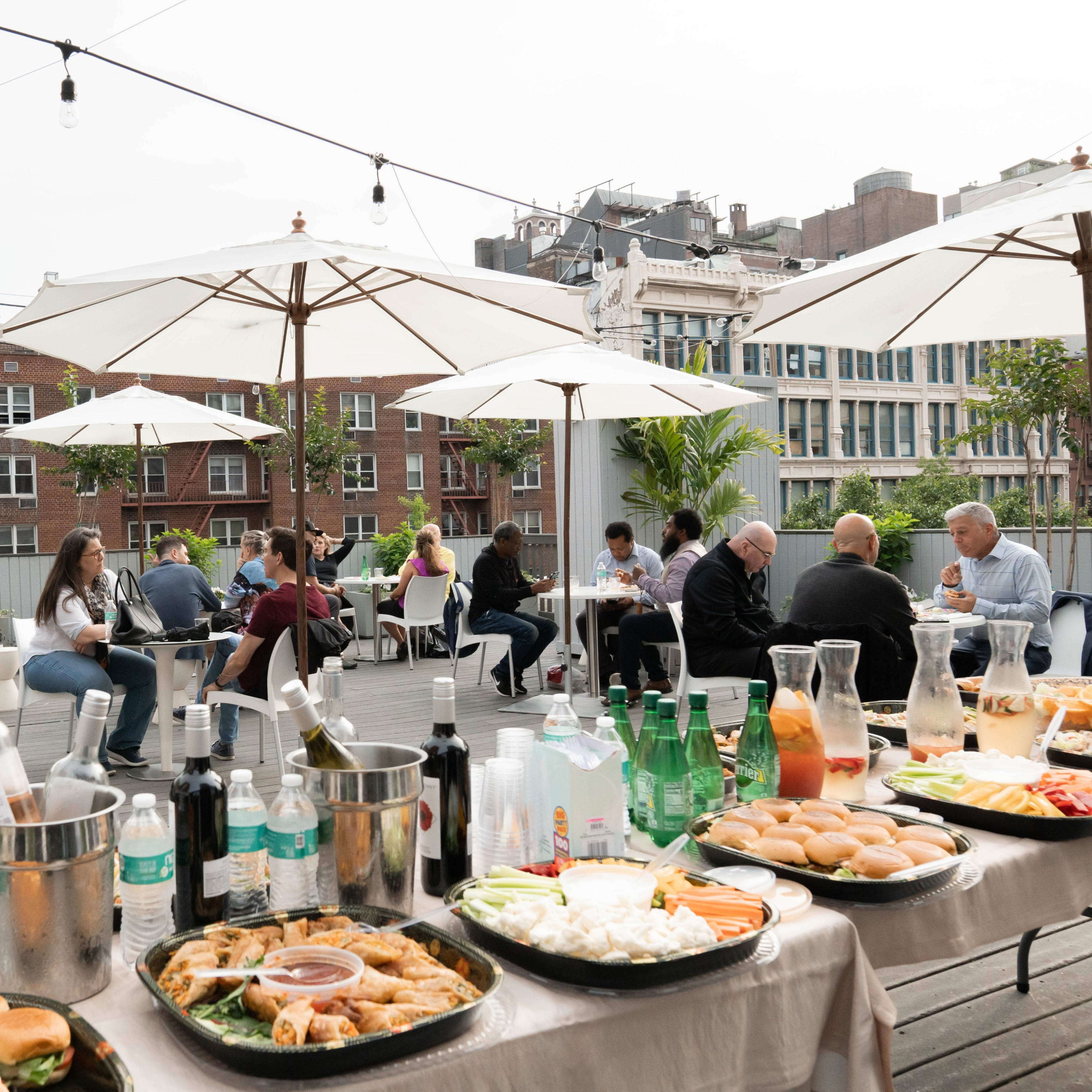 A rooftop gathering features tables with food and drinks, umbrellas for shade, and people socializing in a city setting.