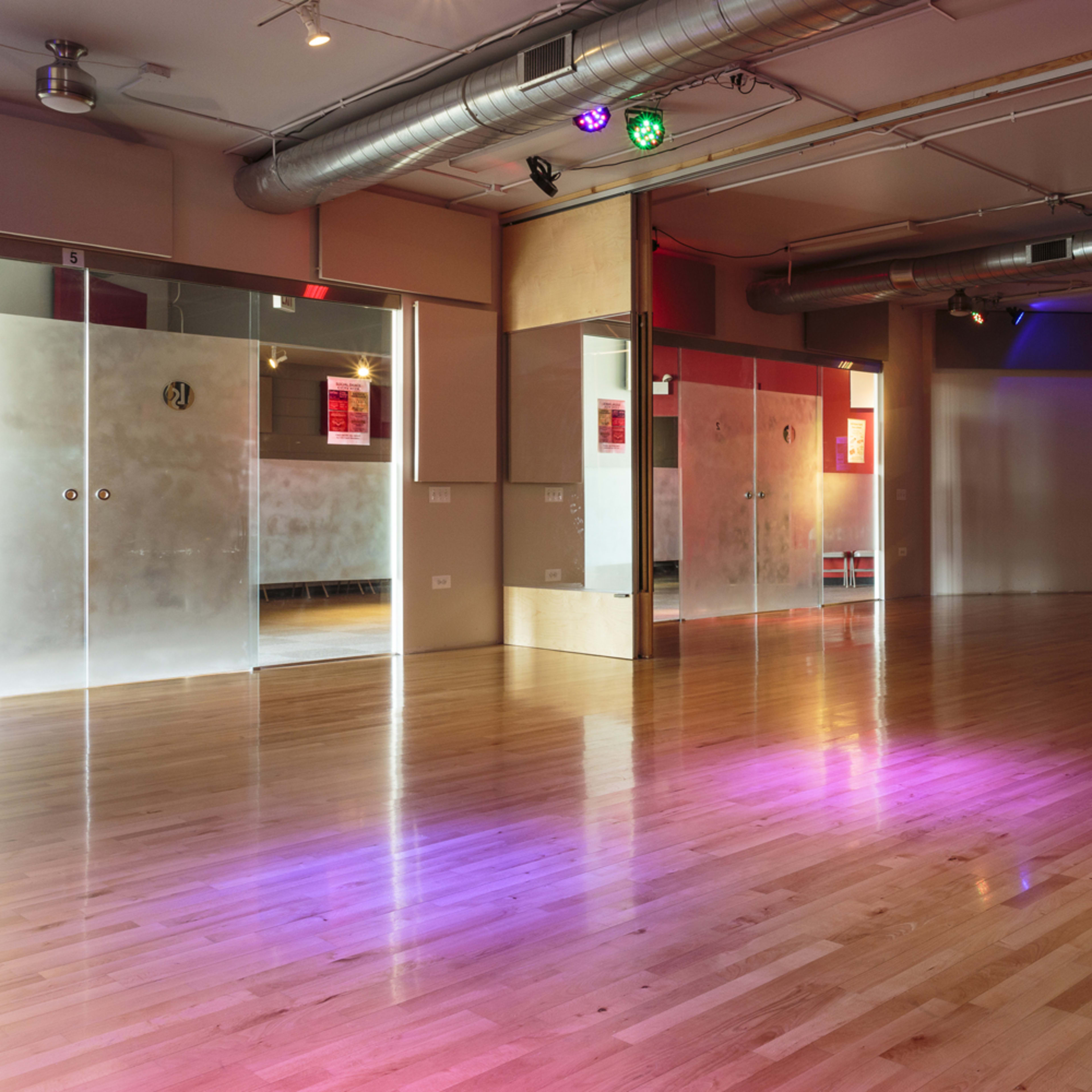 A spacious dance studio with wooden flooring, mirrored walls, and colorful lighting fixtures.