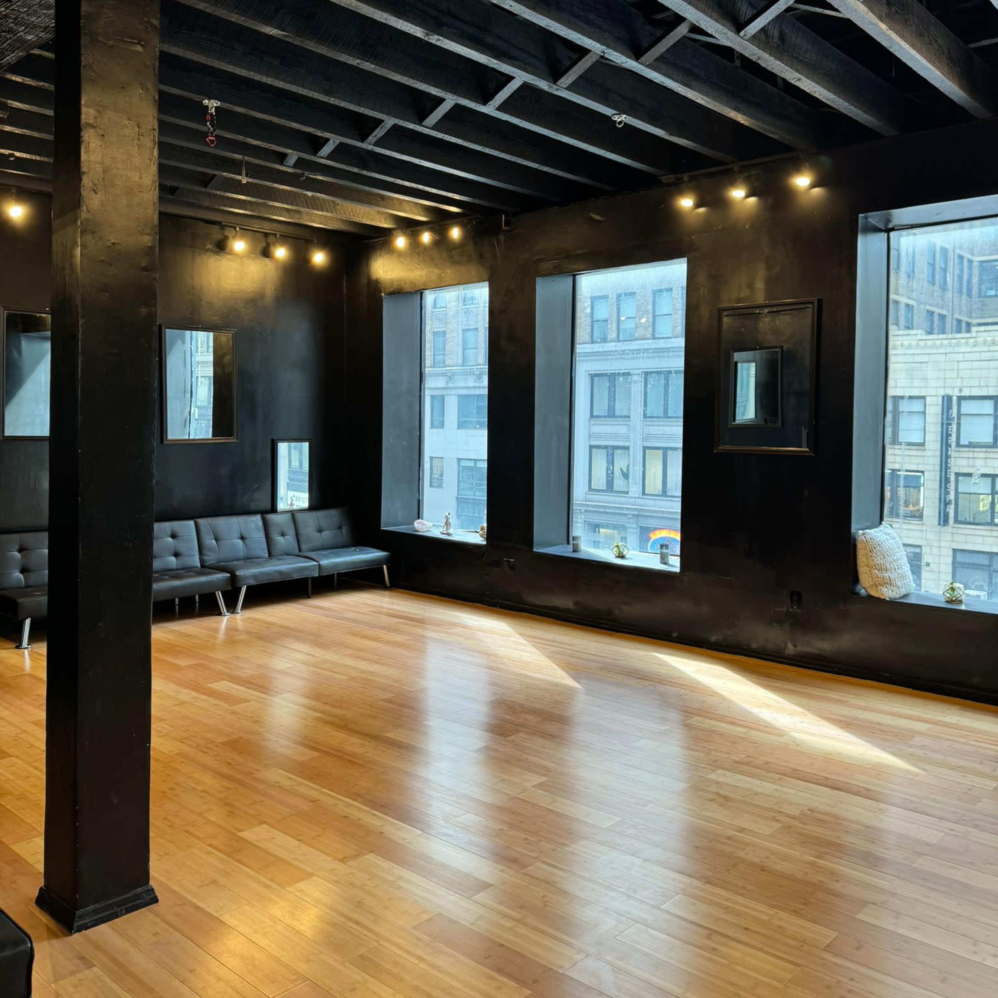An empty room with a hardwood floor, black walls, and large windows allowing light to enter.