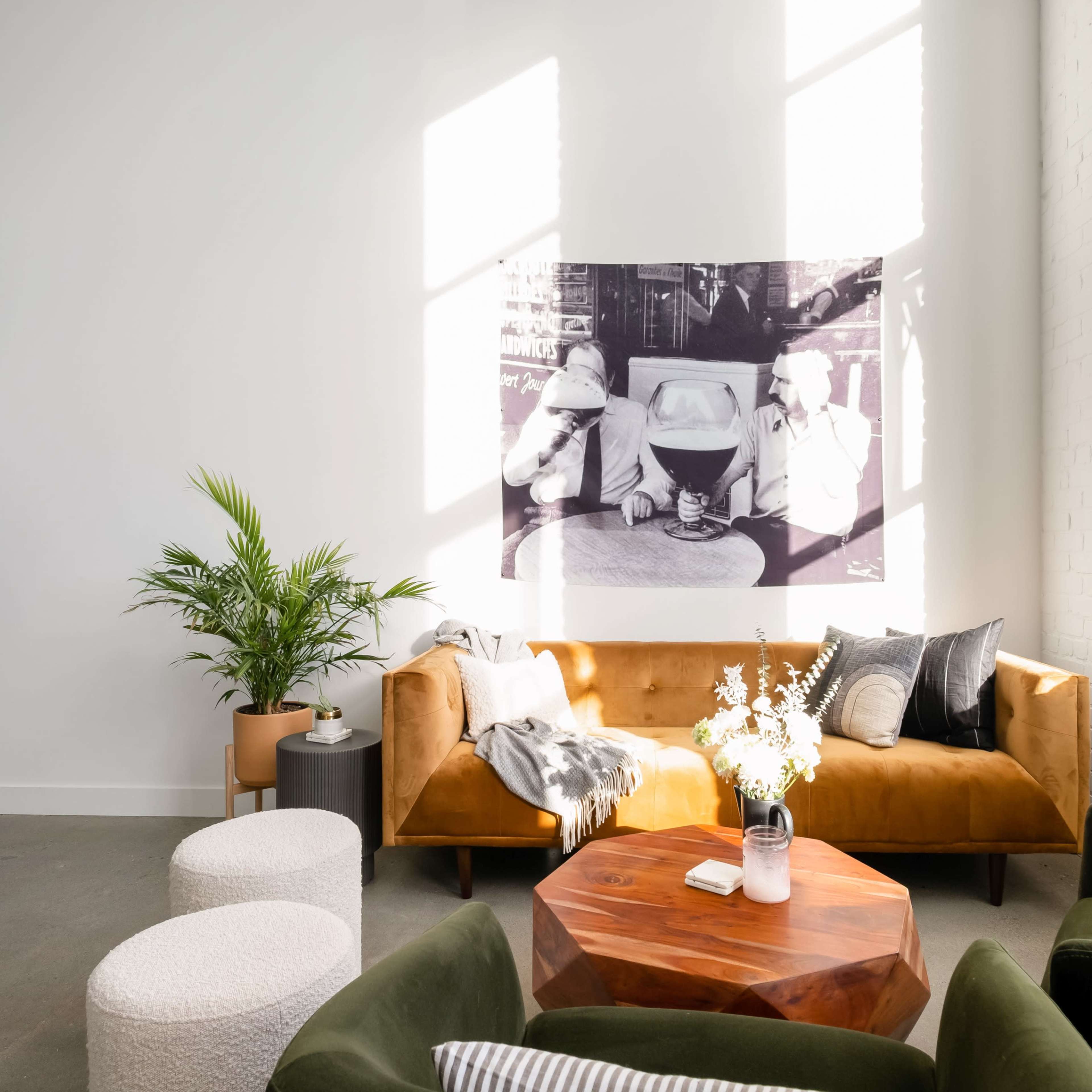 A spacious living room features a brown sofa, a round wooden coffee table, and a large black-and-white photograph on the wall, illuminated by natural light streaming through large windows.