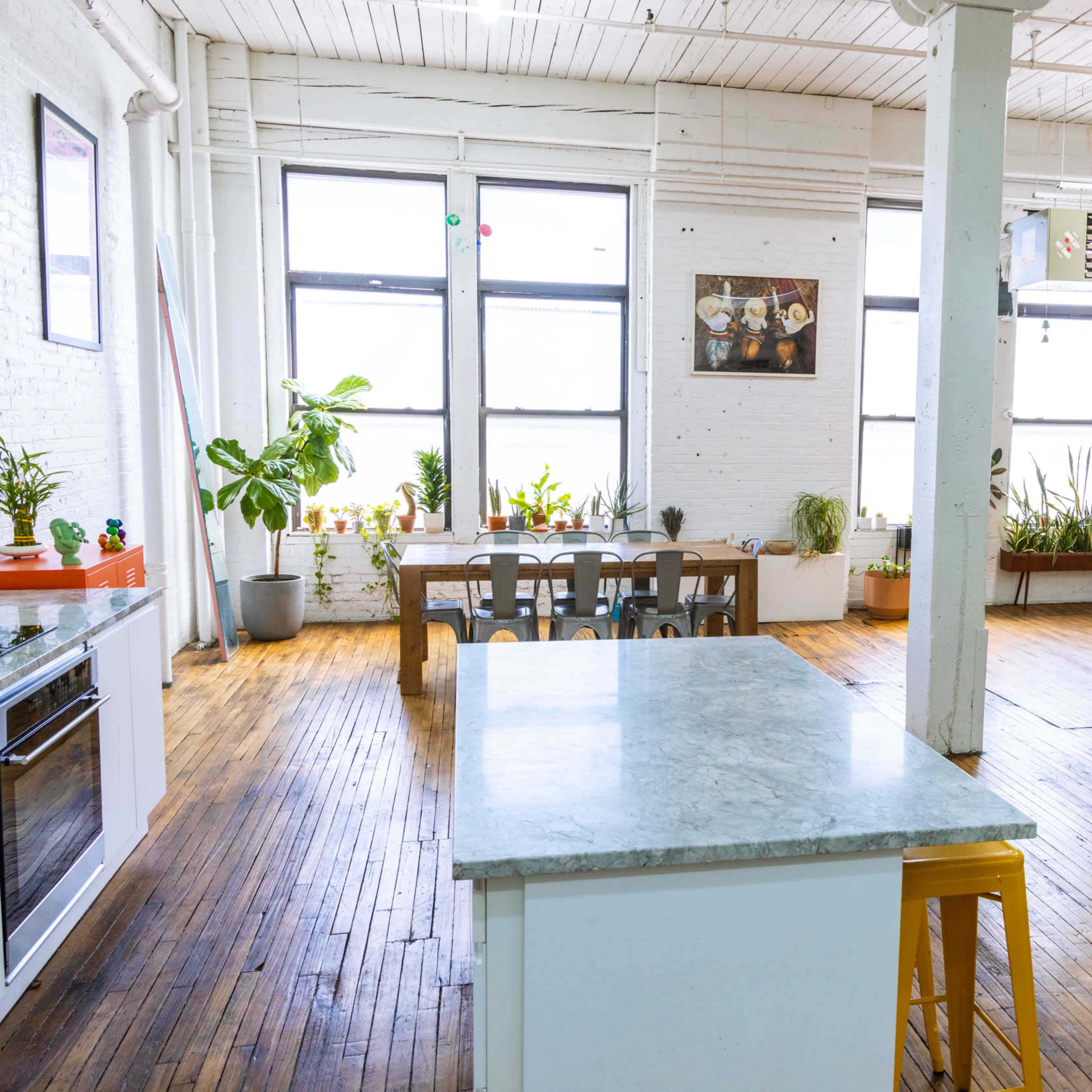 A spacious kitchen and living area with wood flooring, large windows, plants, a dining table, and a leather sofa.