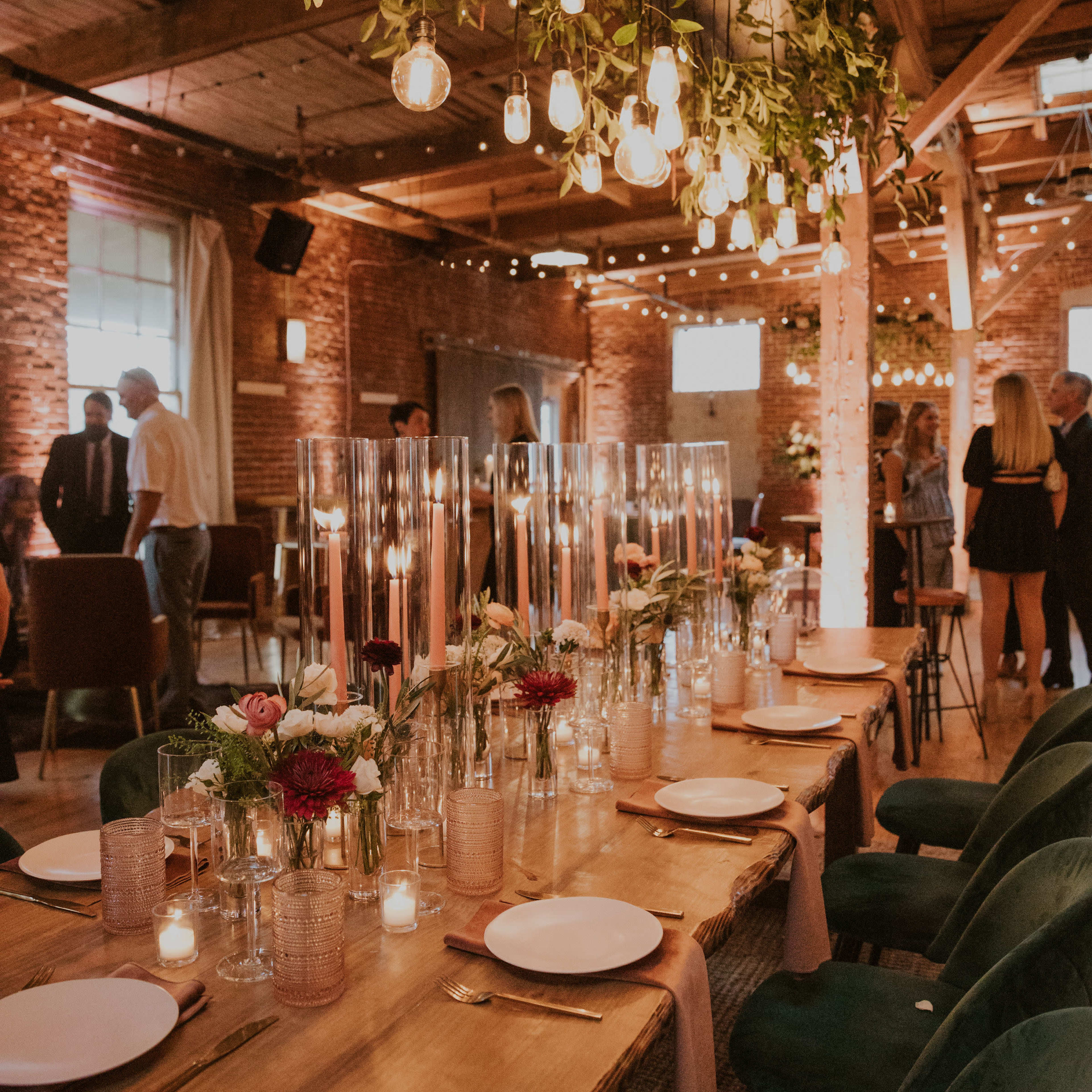 A long wooden dining table is set with candles, plates, and glass vases of flowers under hanging string lights in a rustic event space.