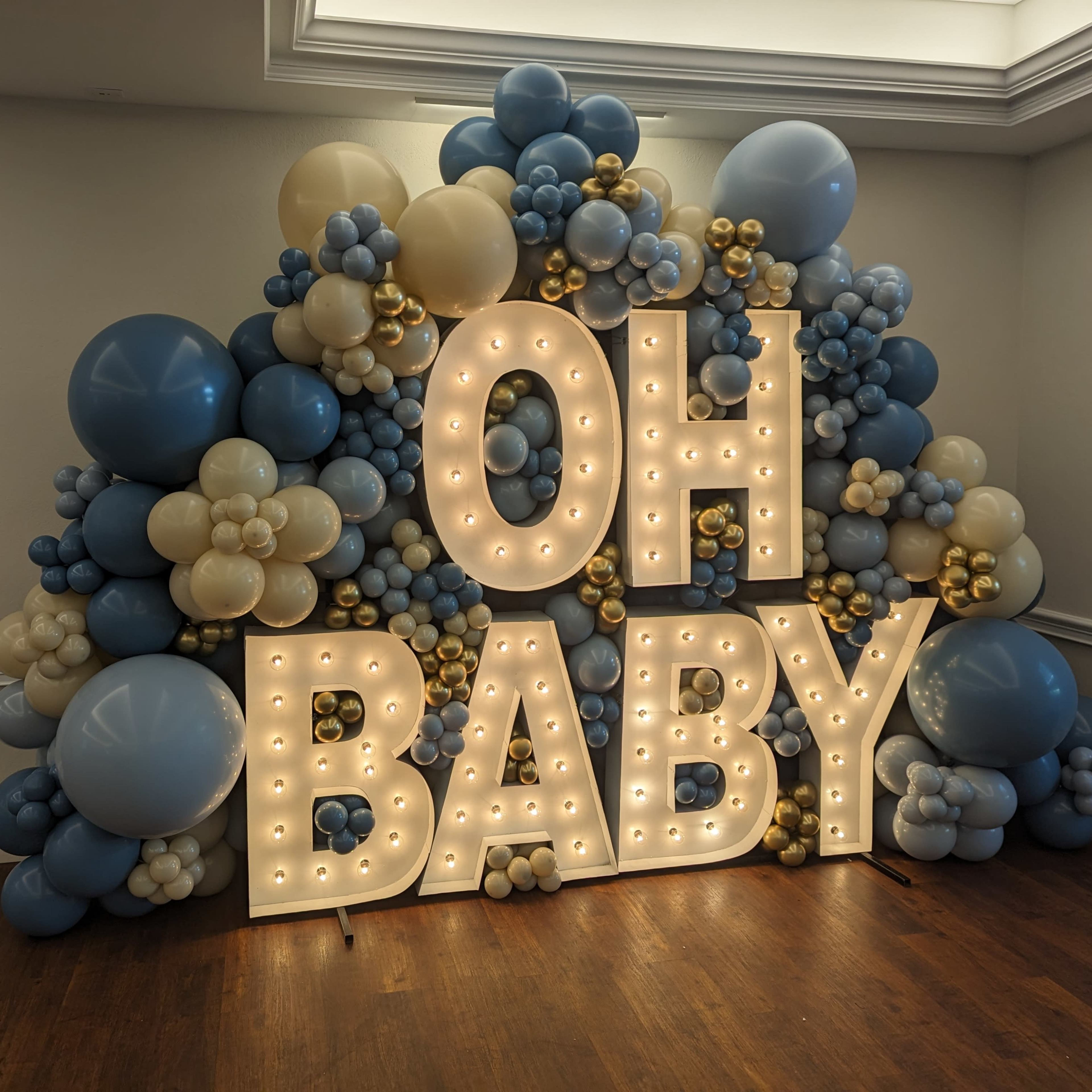 A decorative backdrop made of blue, beige, and gold balloons displays the illuminated phrase "OH BABY."