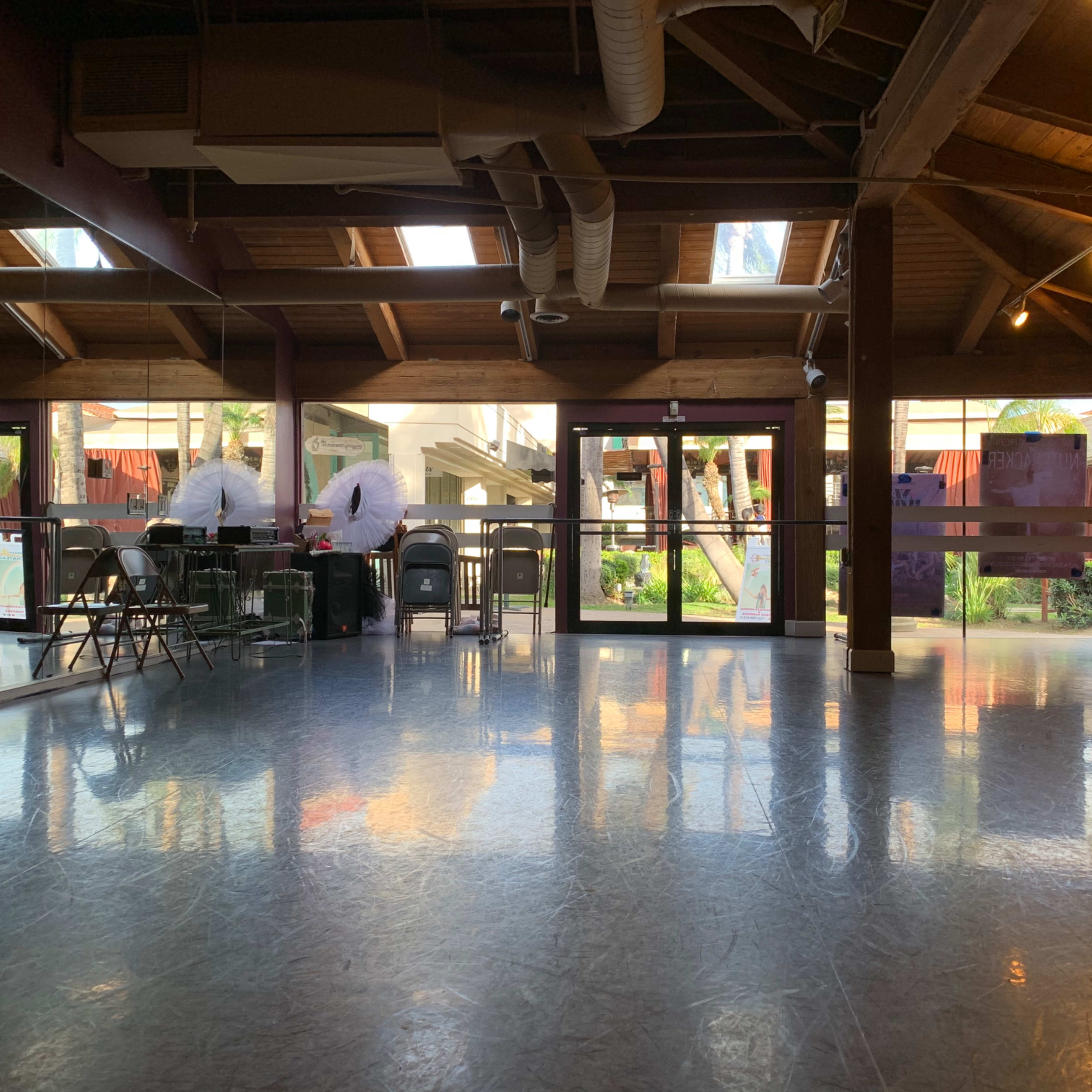 A dance studio with wooden beams and a mirrored wall, featuring empty chairs and large windows that overlook an outdoor area.