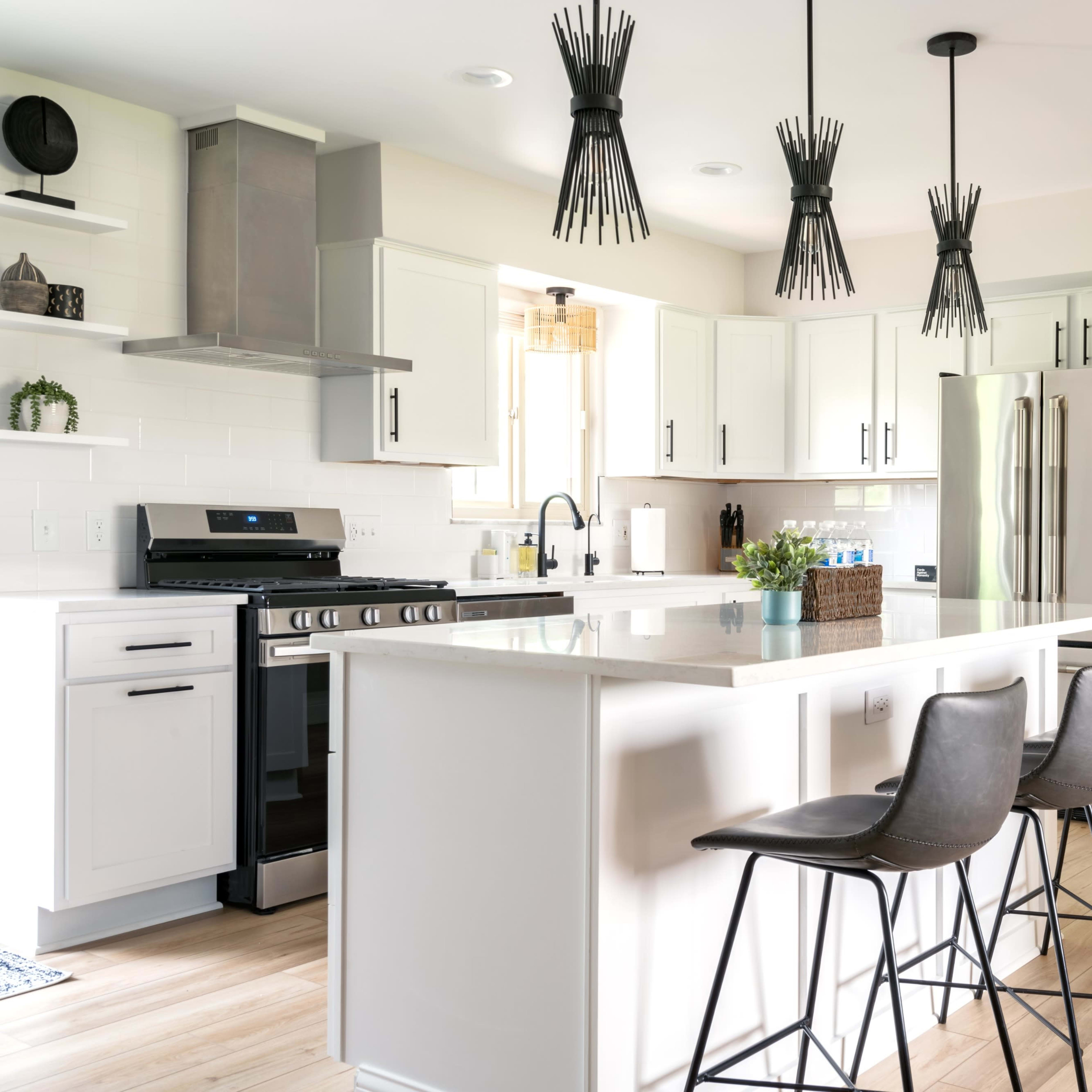 A modern kitchen with a central island, three bar stools, and pendant lighting above, featuring white cabinetry and stainless steel appliances.