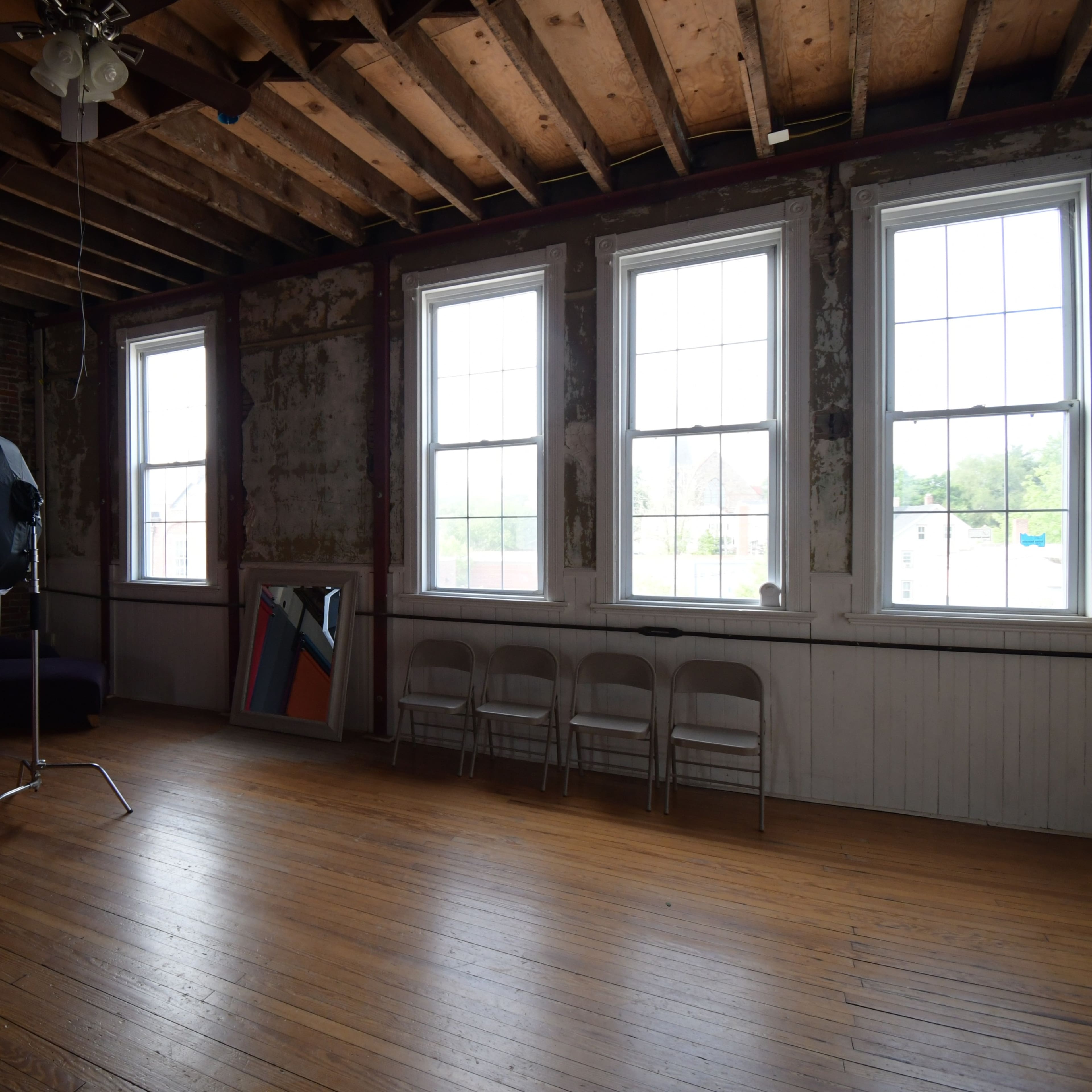 A spacious photo studio with a red backdrop, three tall windows, a mirror, and several white chairs arranged along the wall.