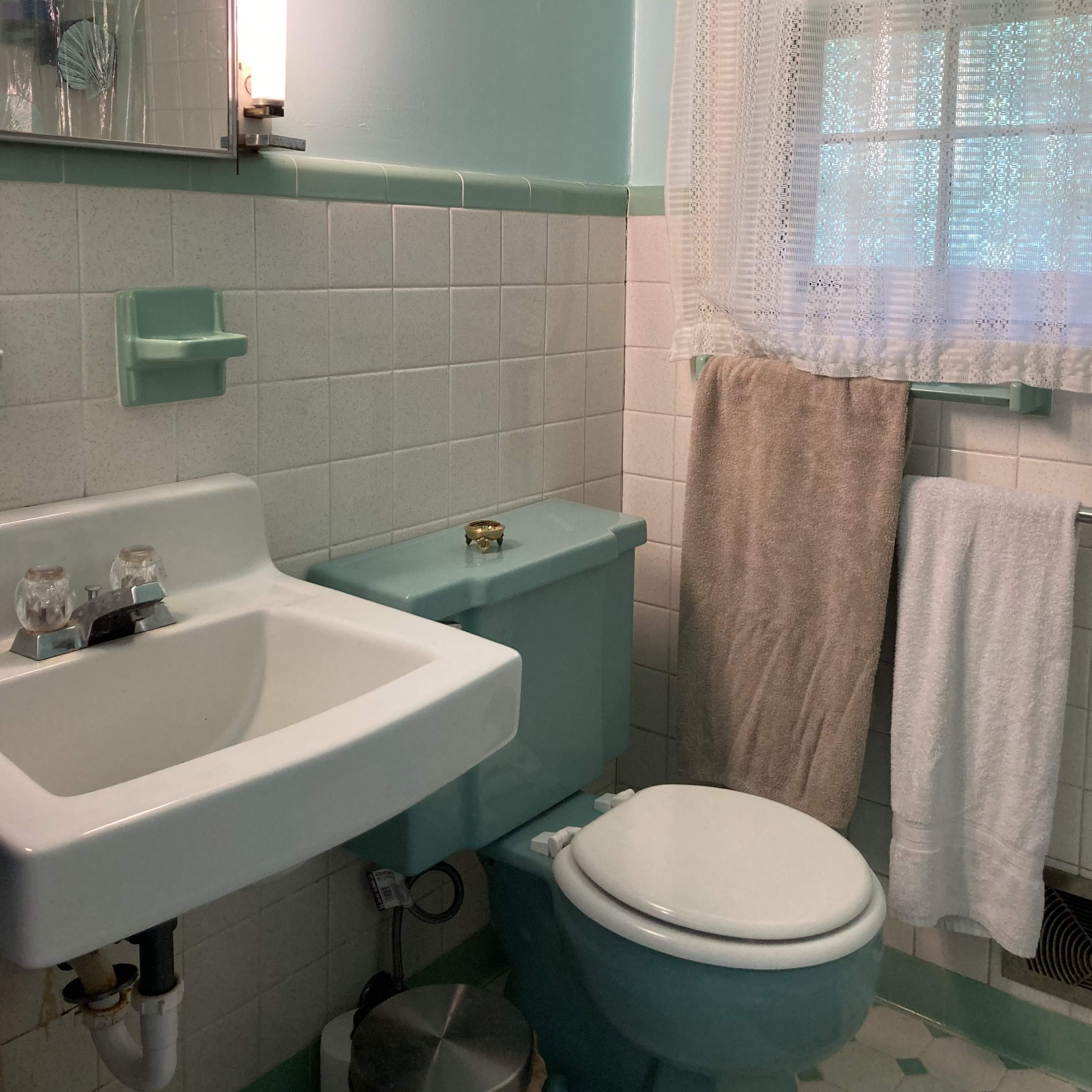 A small bathroom featuring a white toilet, a sink, a mirror with a light, and a window covered by a lace curtain.