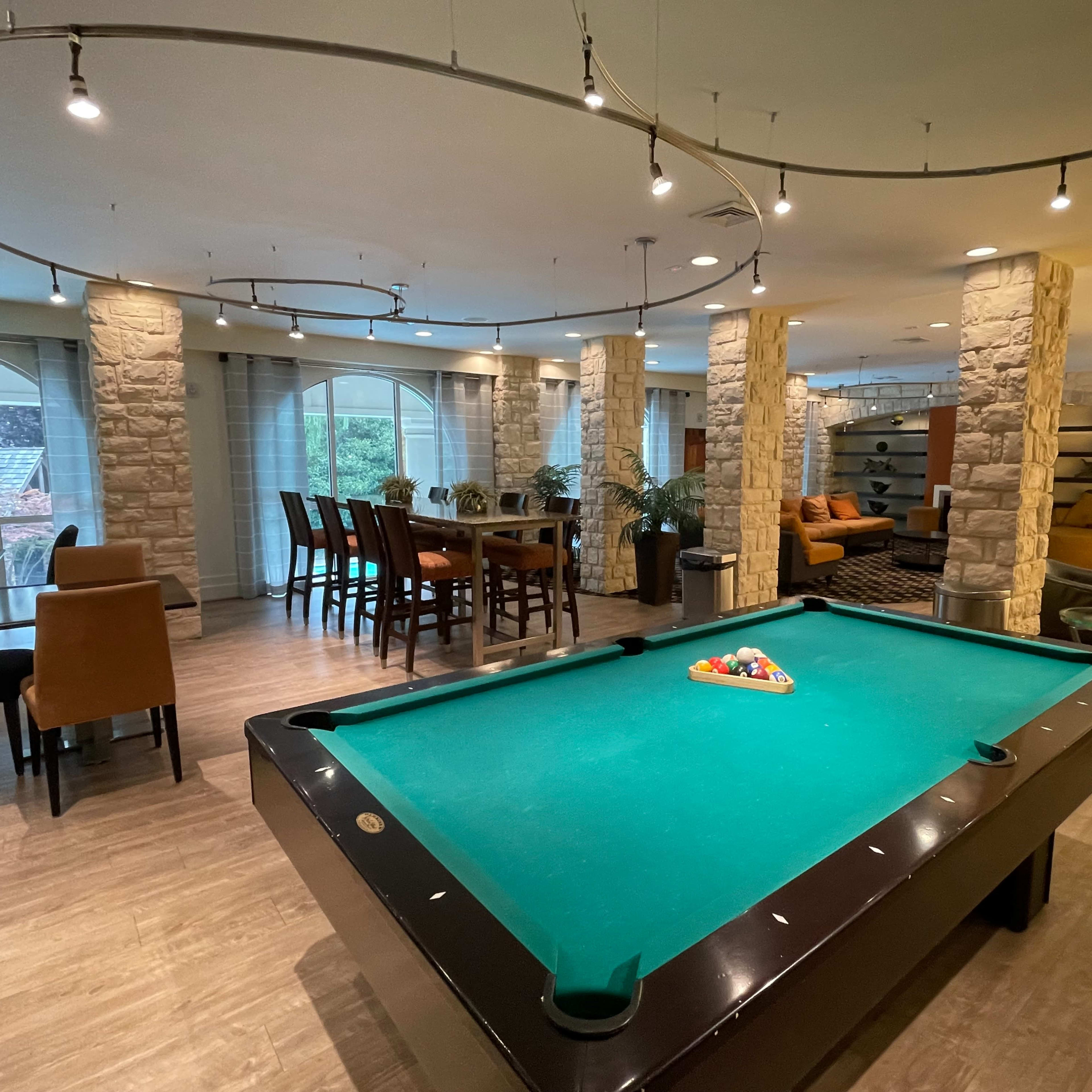 A spacious room features a billiards table at the center, surrounded by seating areas and dining spaces with modern decor.