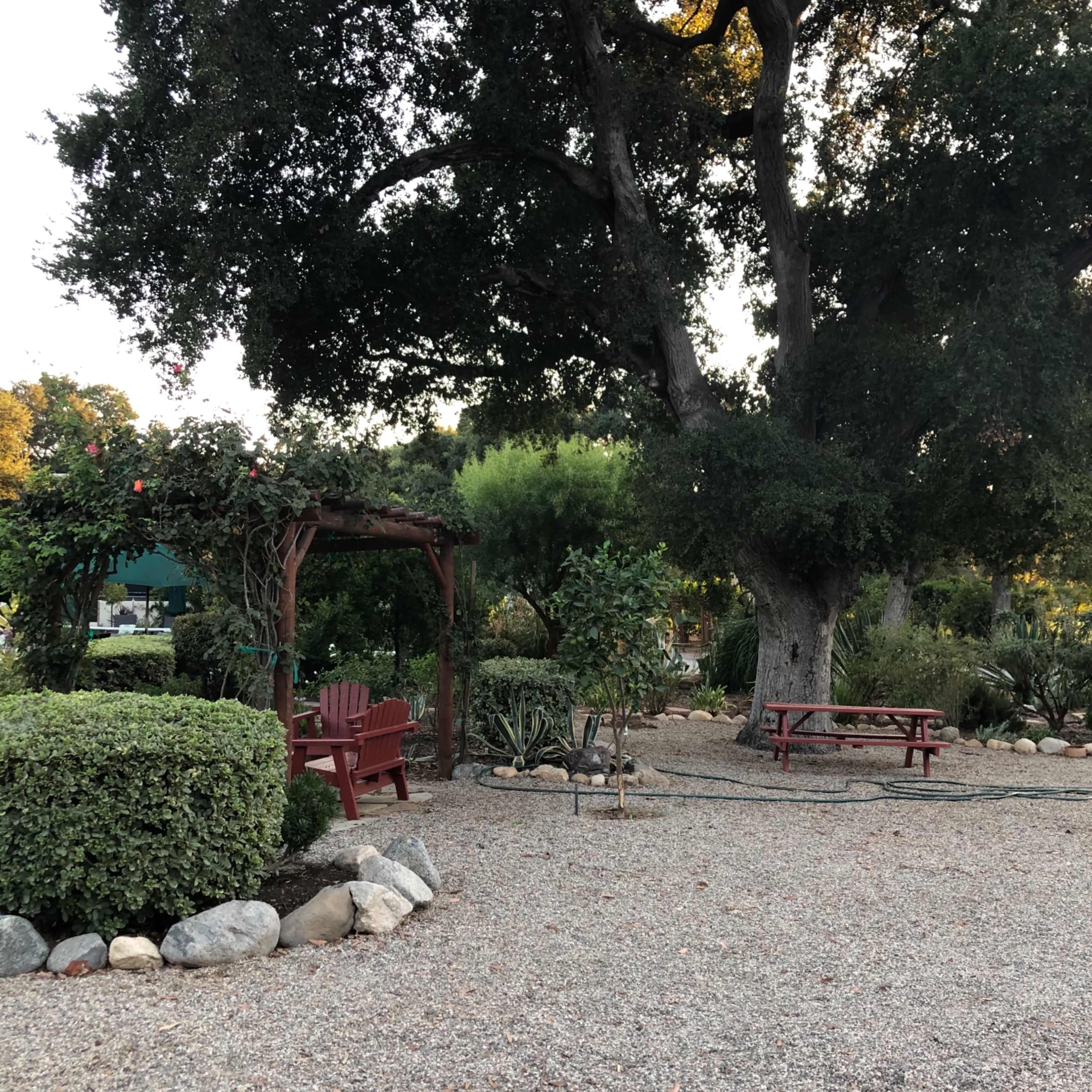 A landscaped garden with a red wooden arbor, chairs, and a picnic table surrounded by trees and shrubbery.