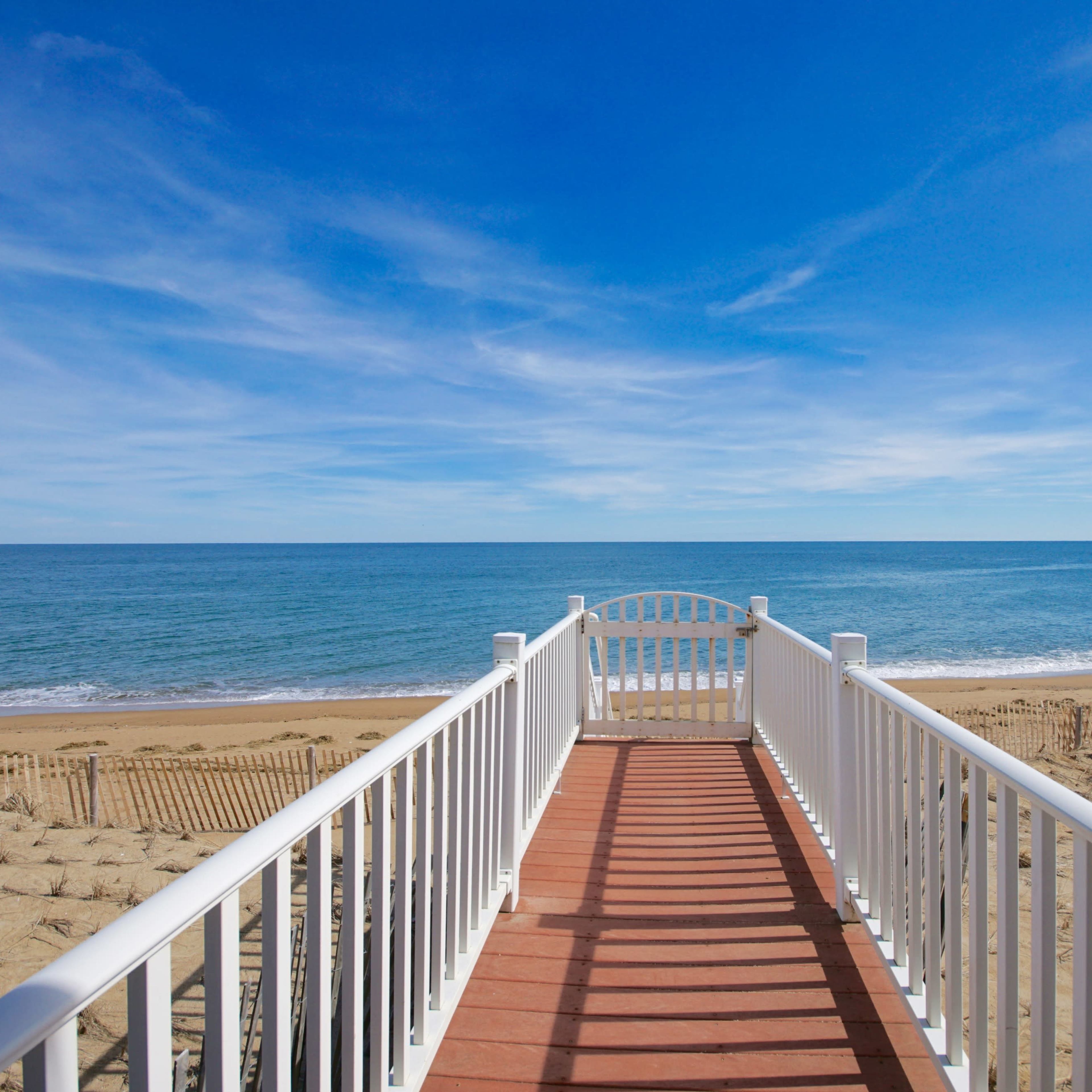A white wooden walkway extends towards a sandy beach and calm ocean under a clear blue sky.
