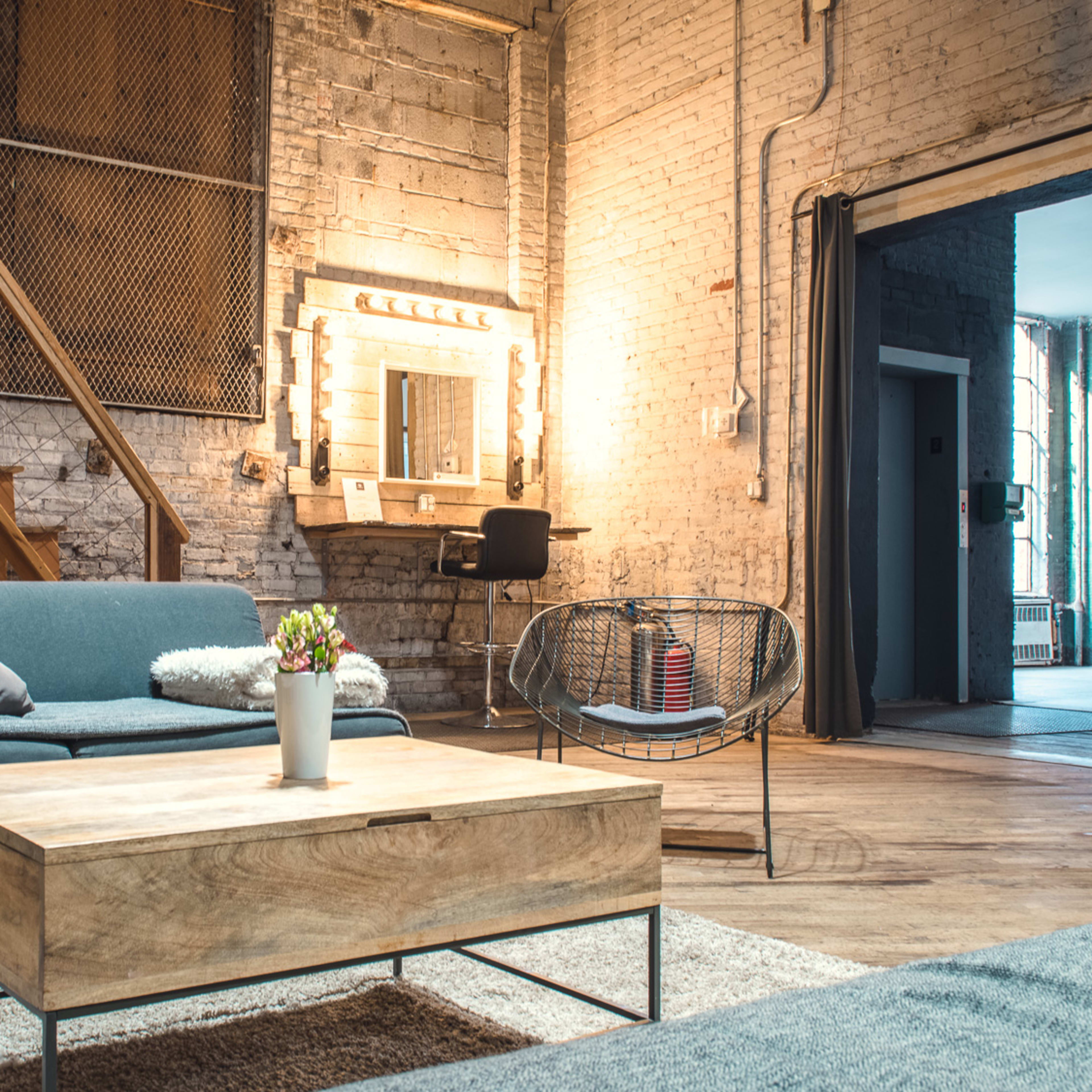 A modern, industrial-style living space with a blue sofa, a wooden coffee table, and a mirrored vanity, set against exposed brick walls and wooden flooring.