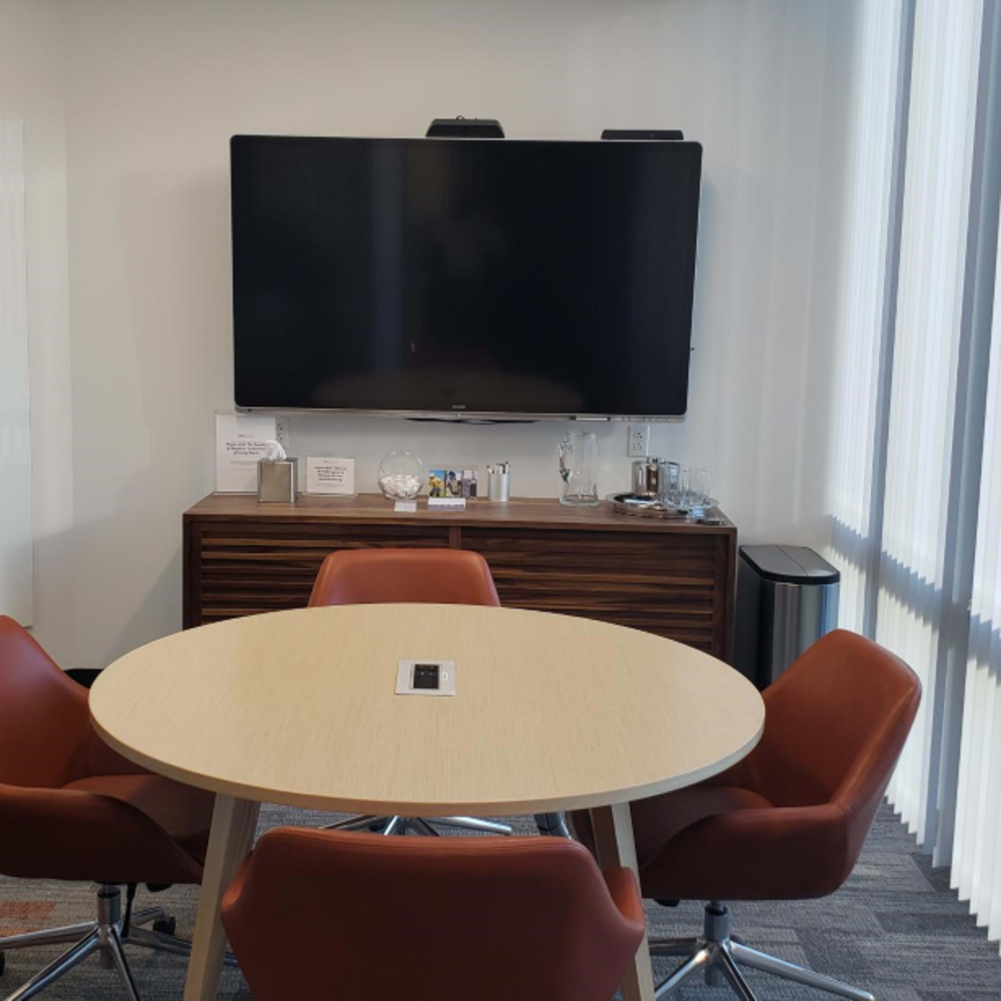 A modern conference room features a round table surrounded by four leather chairs, a wall-mounted television, and a wooden cabinet.