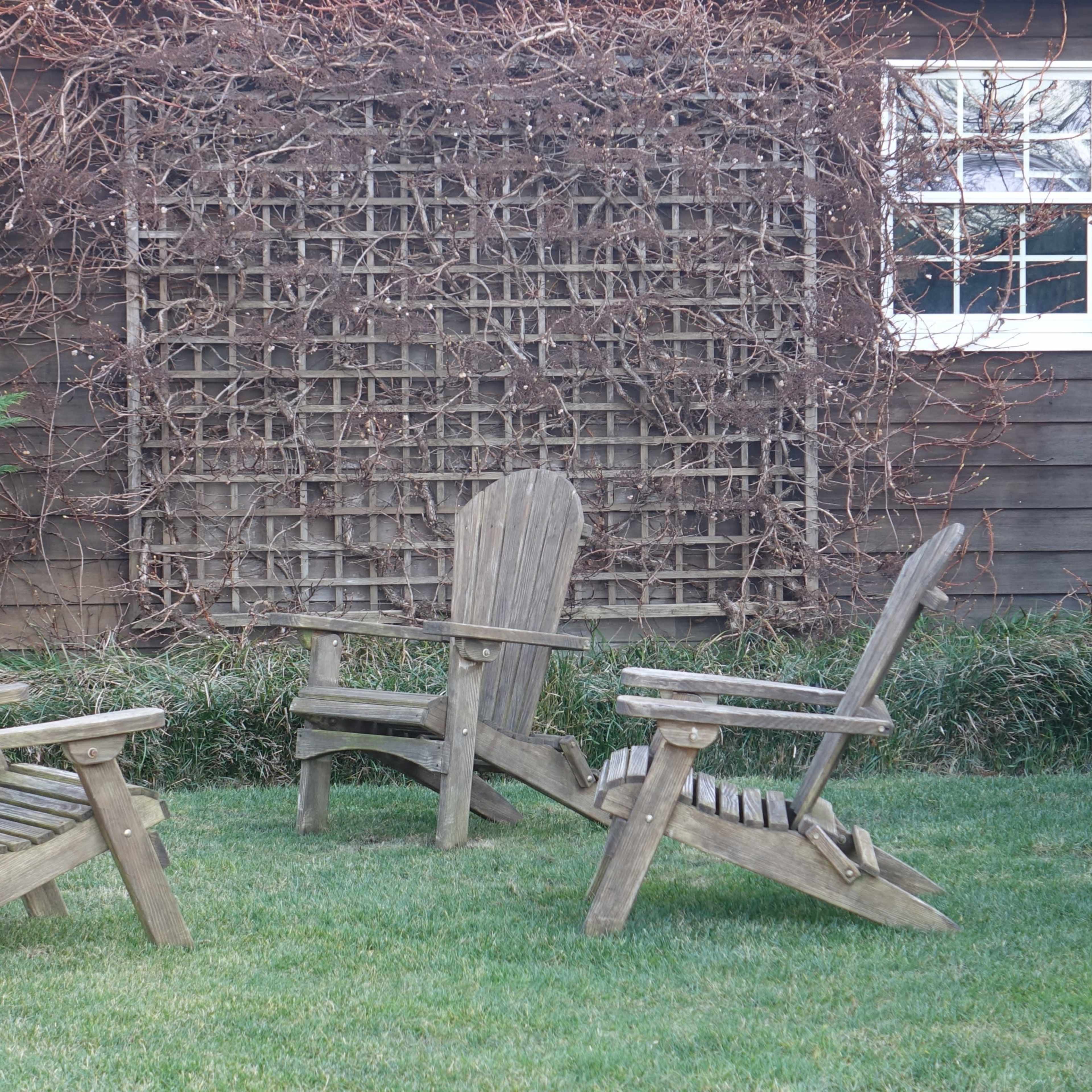 A set of wooden Adirondack chairs is arranged on a grassy lawn next to a wooden wall covered with a trellis of vines.