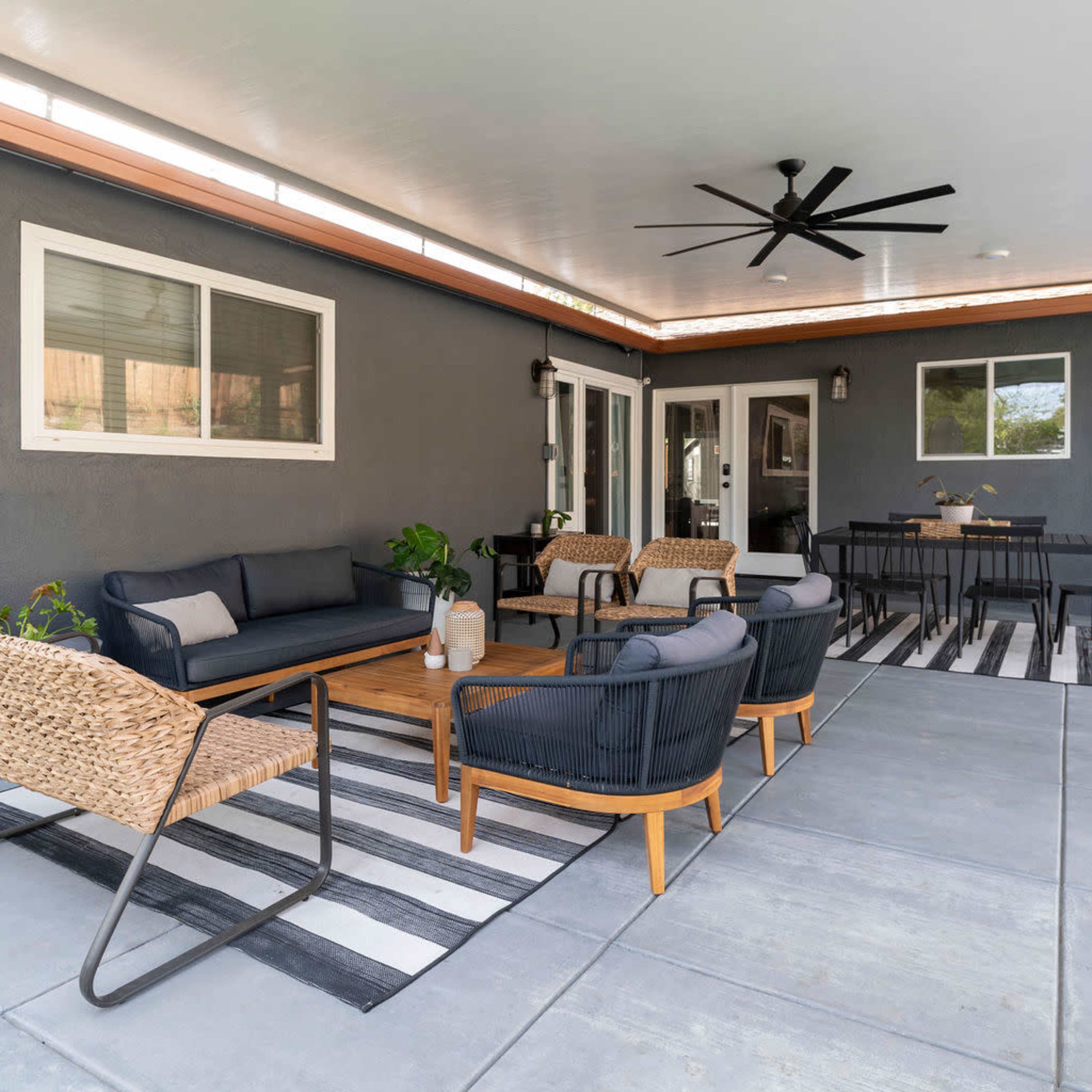A covered outdoor patio features a seating area with black wicker chairs, a wooden coffee table, and a dining space in the background.
