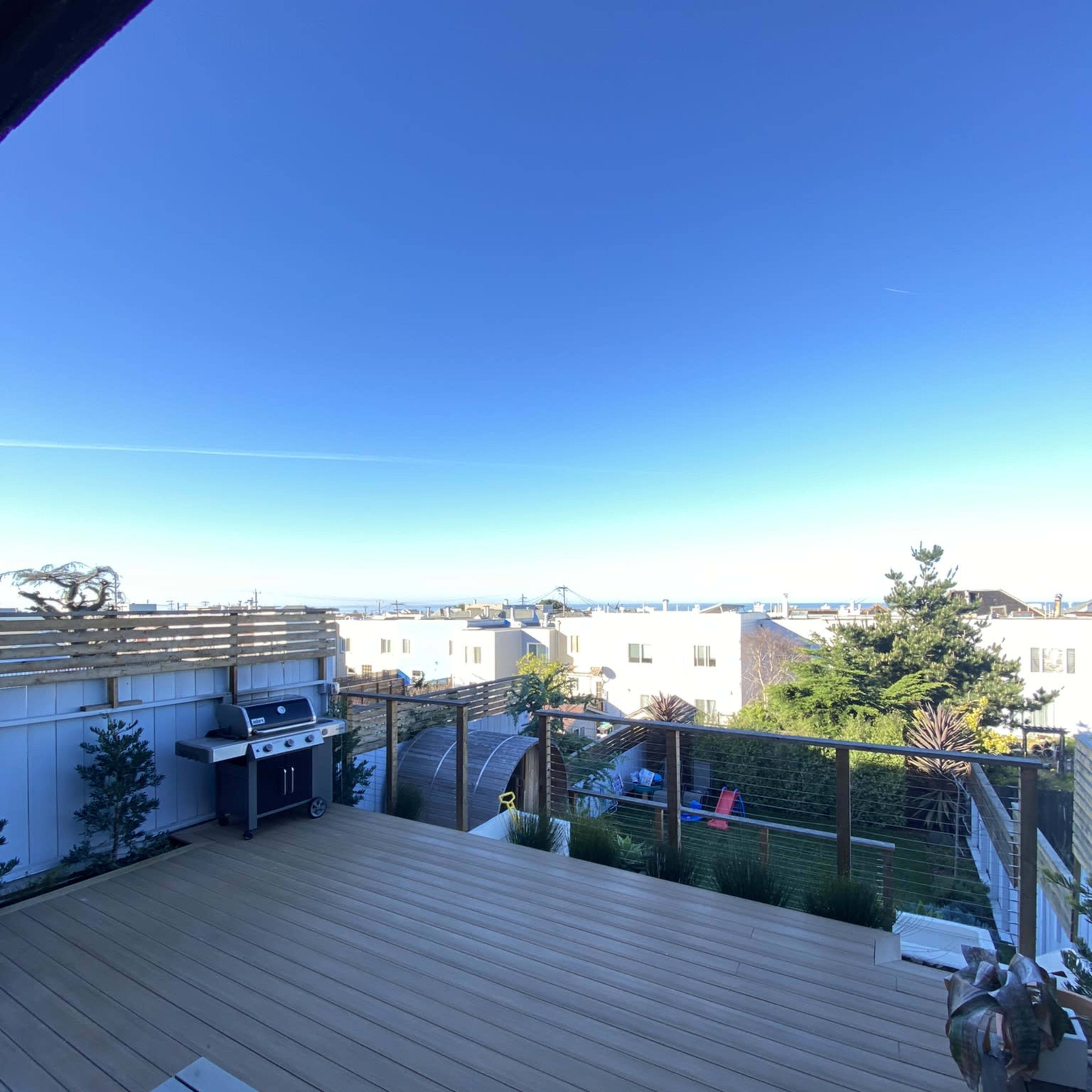 A wooden deck overlooking a landscape with houses and the ocean in the distance under a clear blue sky.