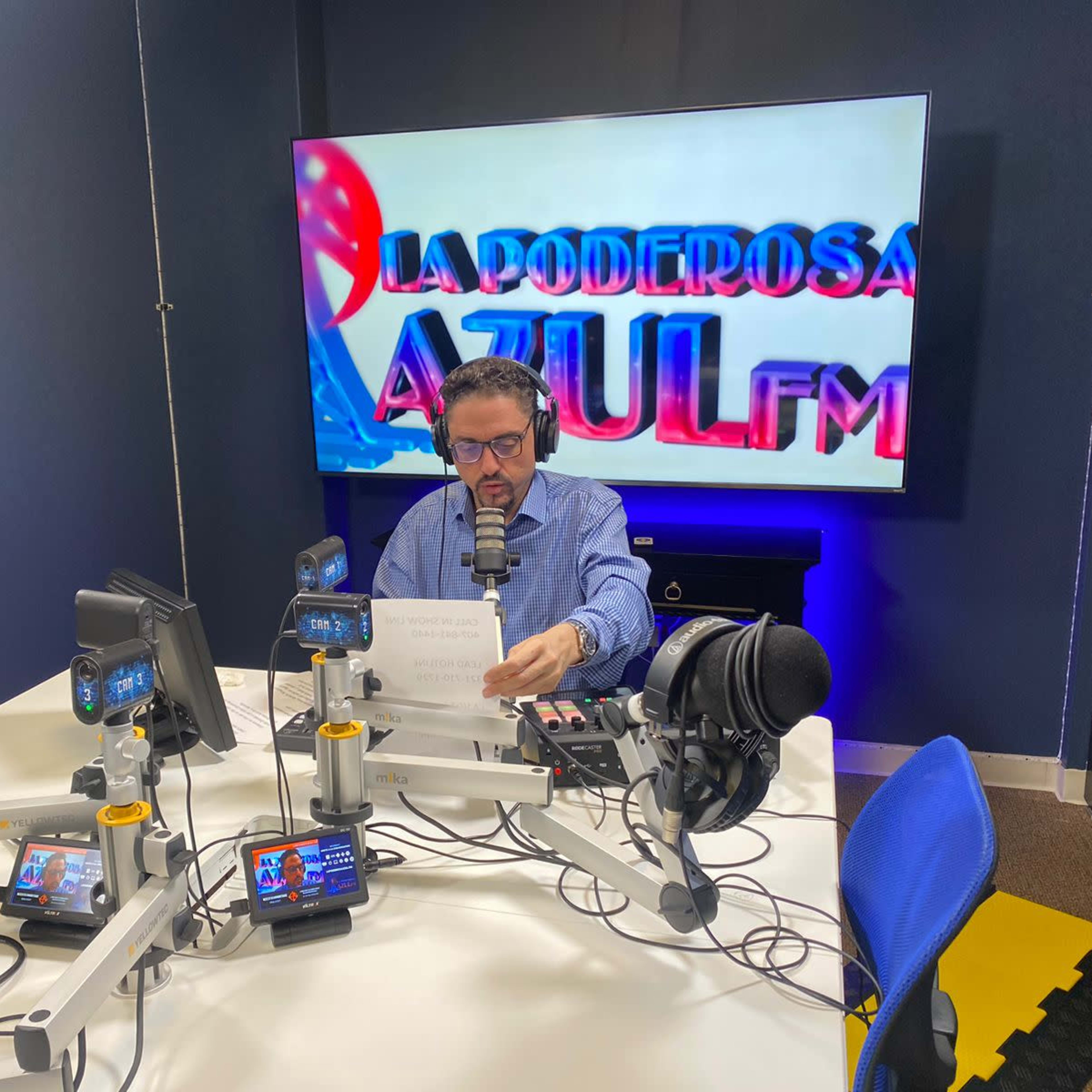 A man wearing headphones reads from a paper while seated at a radio broadcast table with microphones and screens, in front of a large display that says "LA PODEROSA AZUL FM."