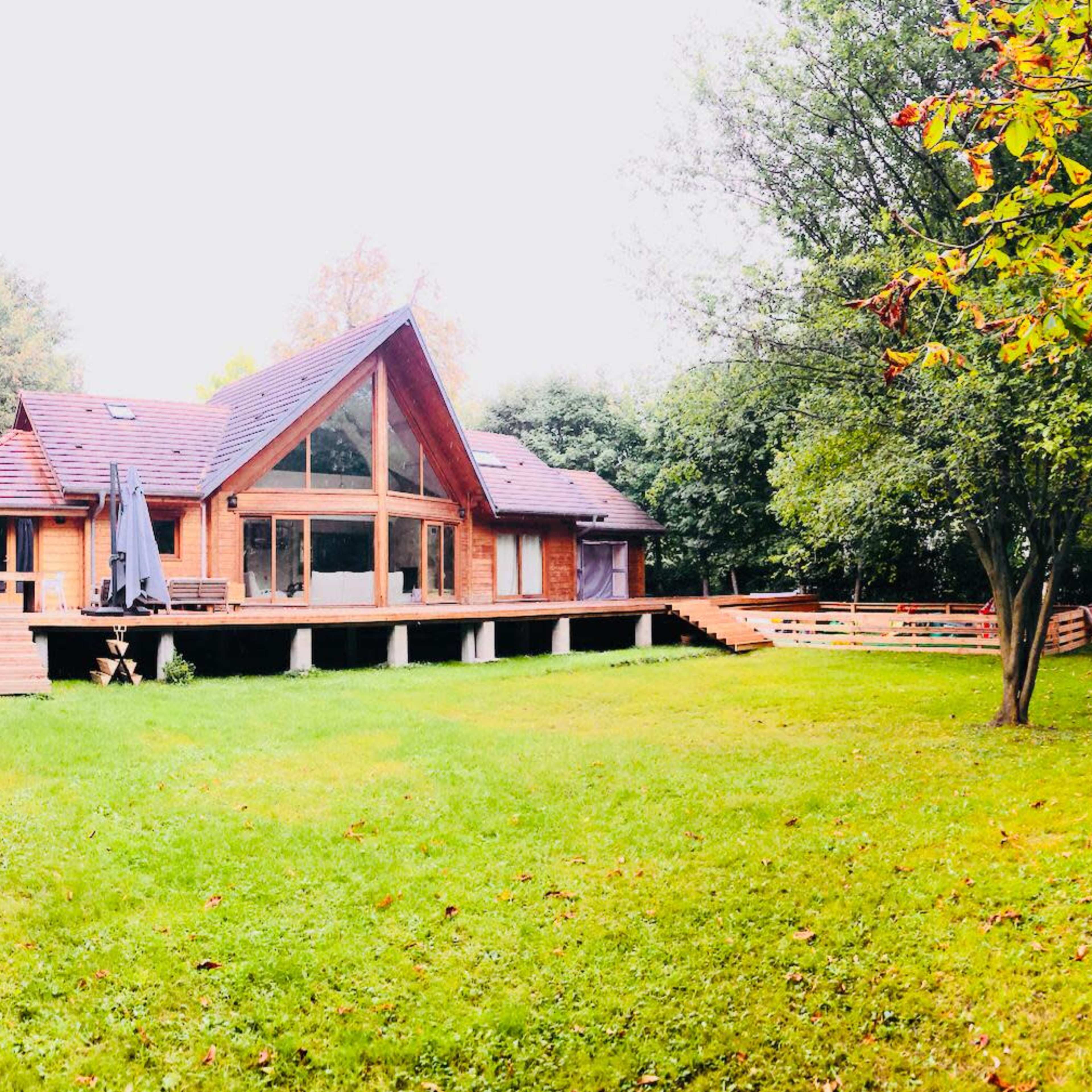A wooden house with a triangular roof and large windows sits on a grassy lawn surrounded by trees.
