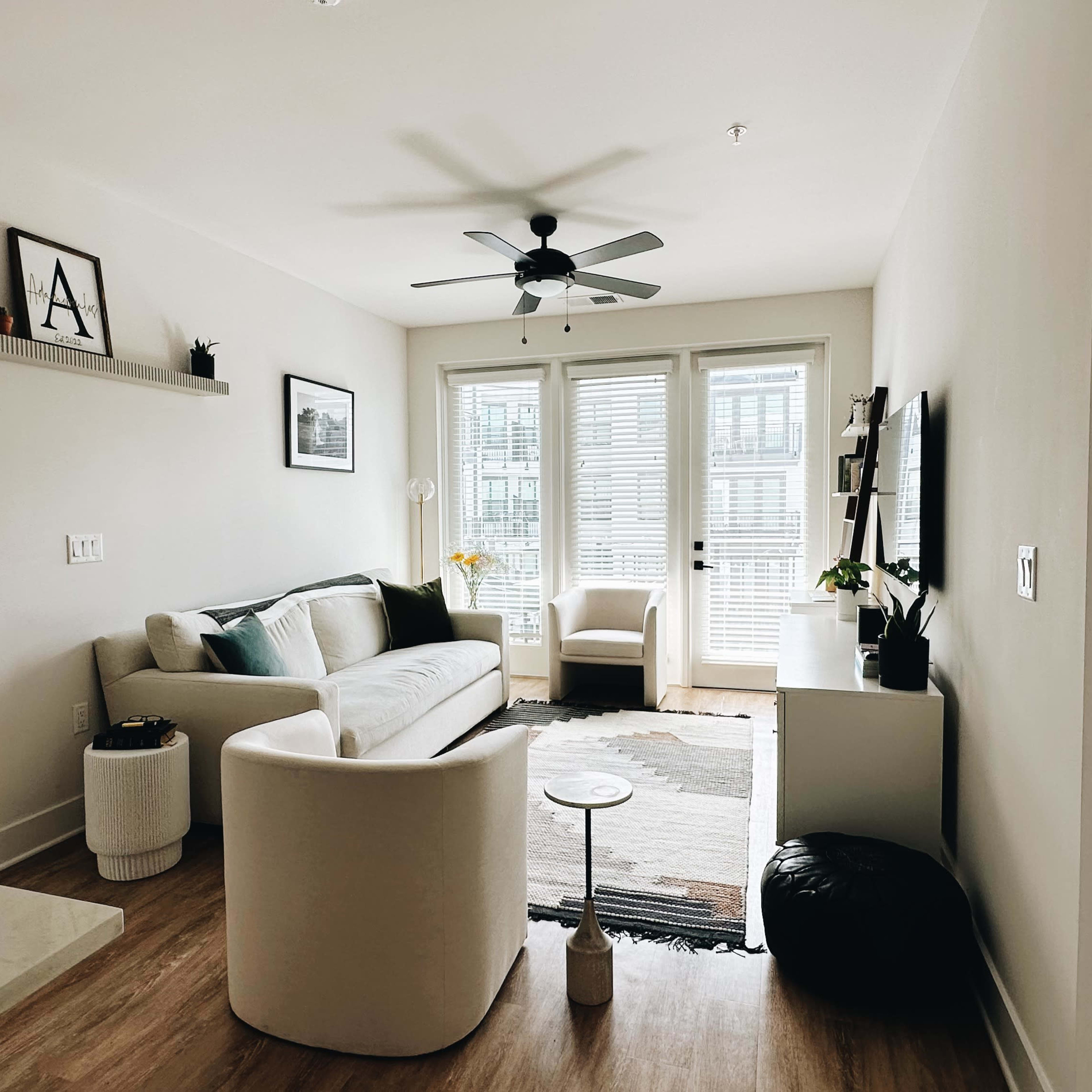 A bright living room features a light-colored sofa, a white accent table, and large windows with blinds, leading to a balcony.