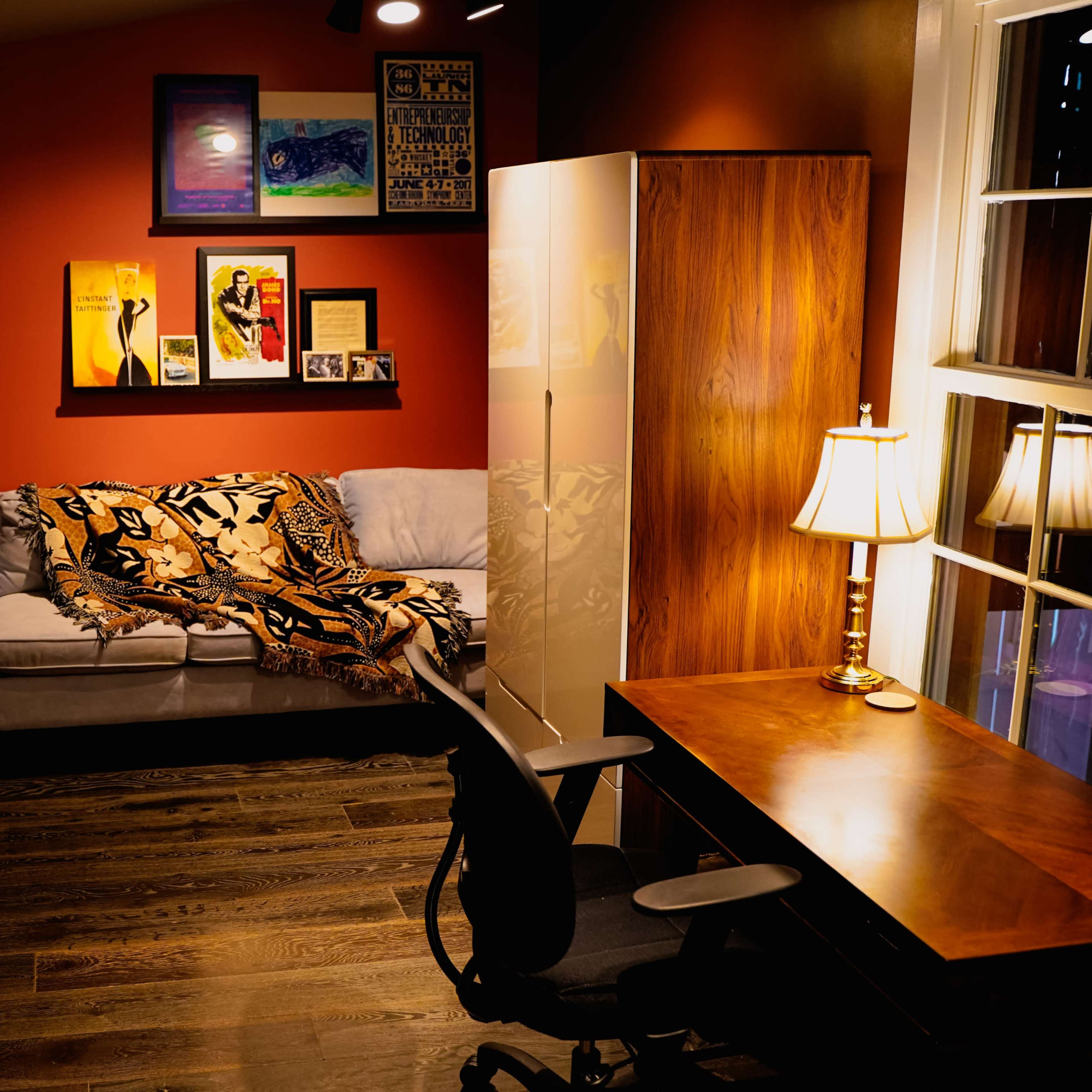 The room features a sofa with a blanket, a desk and chair, a bookshelf filled with books, and various framed artworks on the walls.