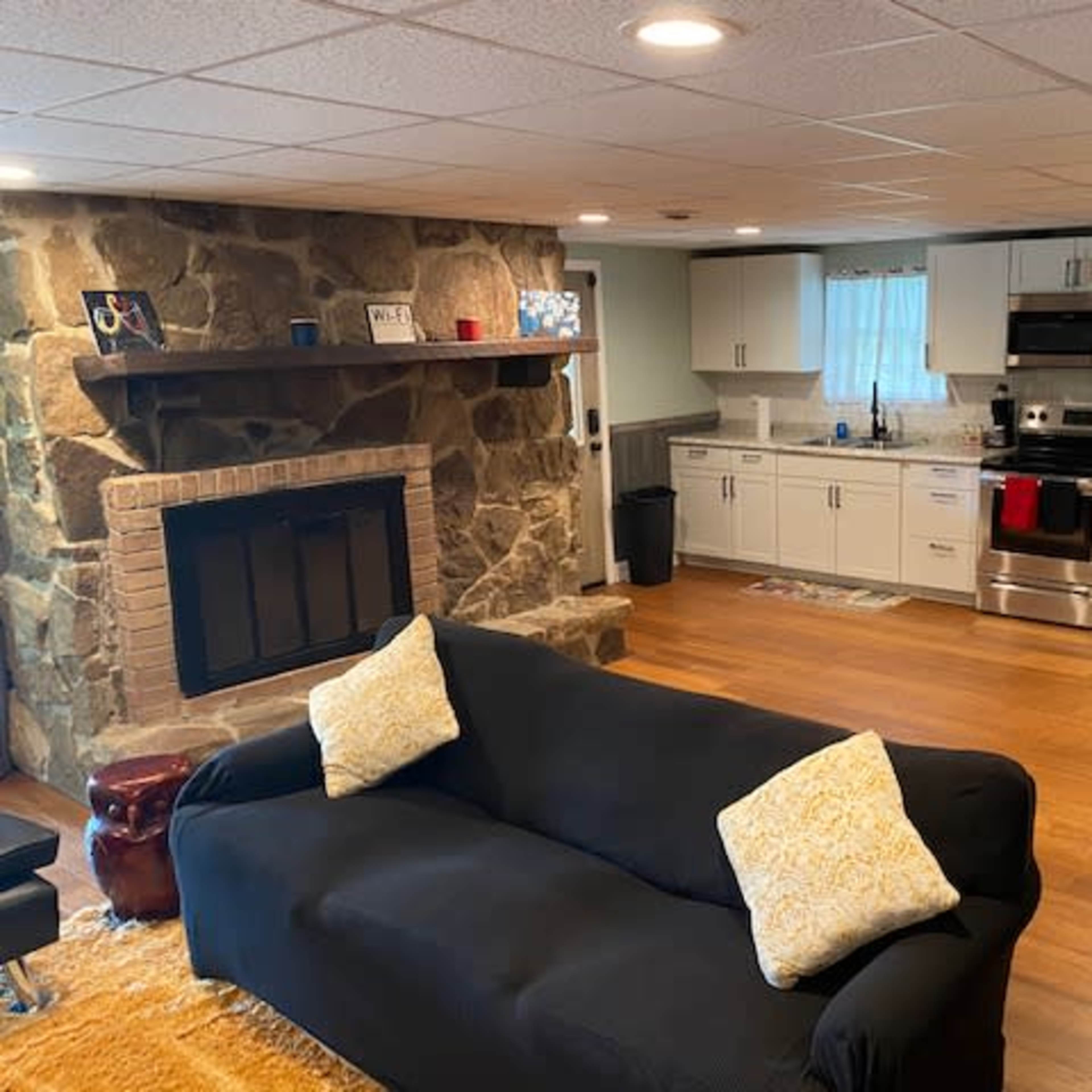 A cozy living space with a black couch, a stone fireplace, and an adjacent kitchen featuring stainless steel appliances and white cabinets.