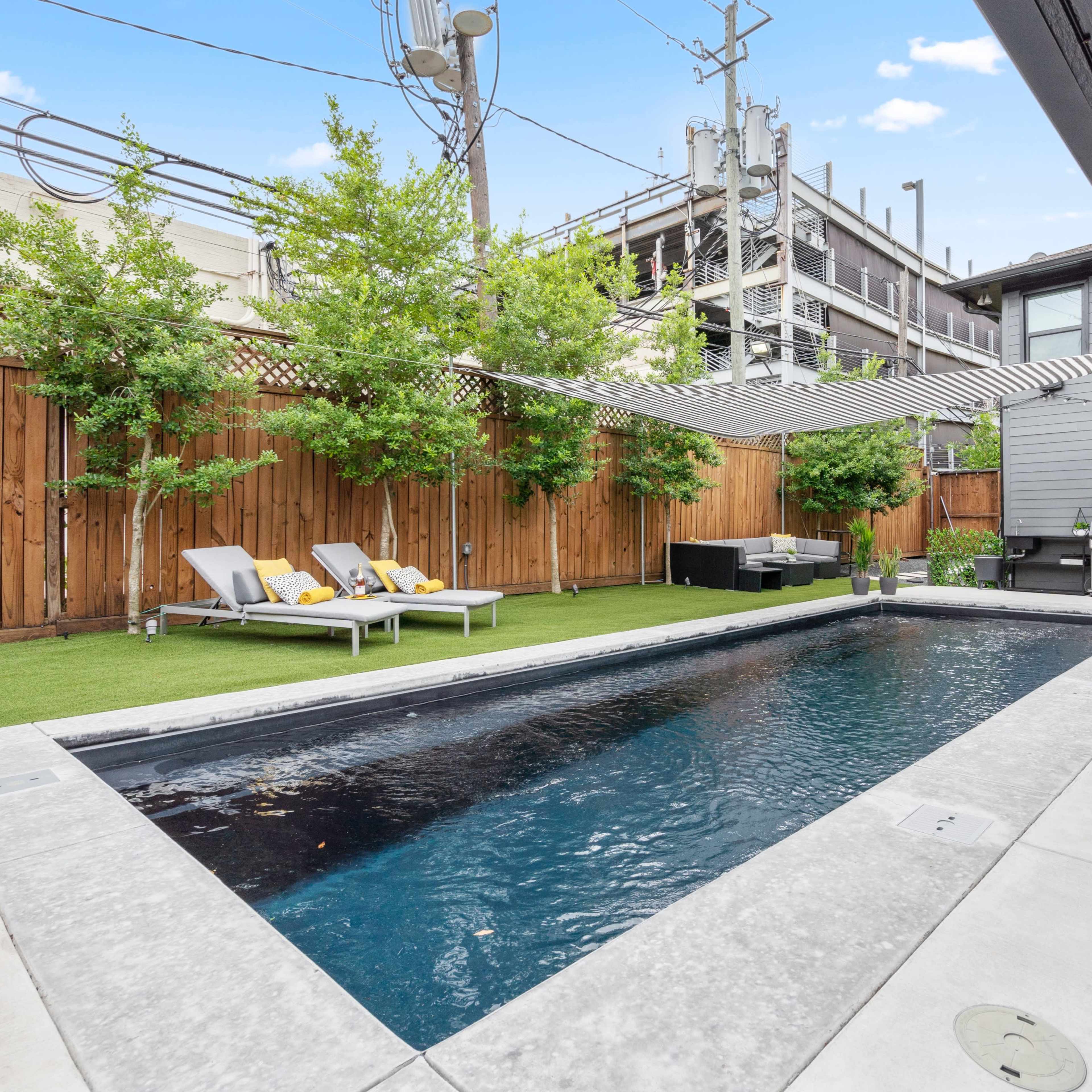 A modern backyard featuring a rectangular swimming pool, lounge chairs, and a small outdoor kitchen area, surrounded by wooden fencing and greenery.