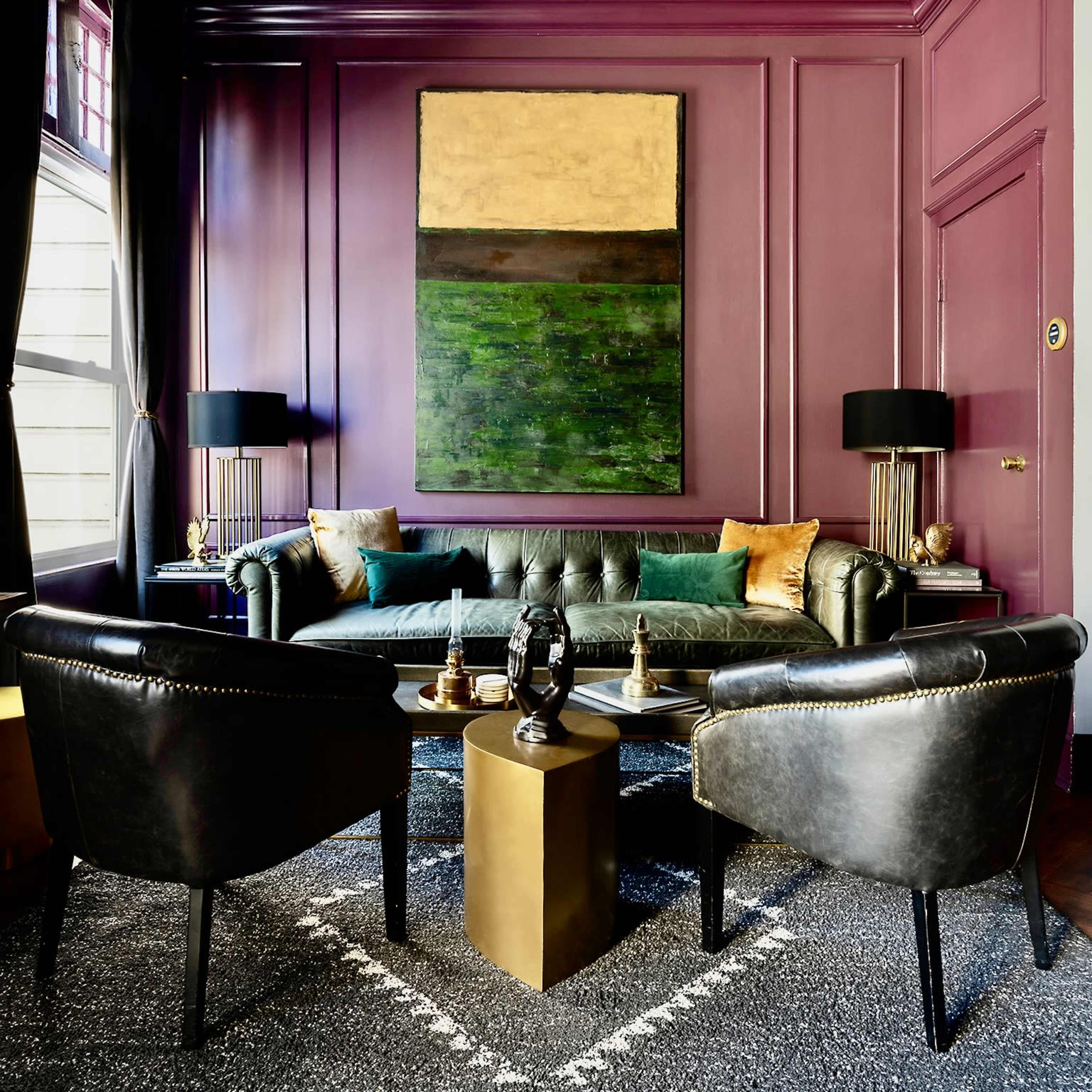 A stylish sitting area with a green leather couch, two black chairs, and a large abstract painting on a purple wall.