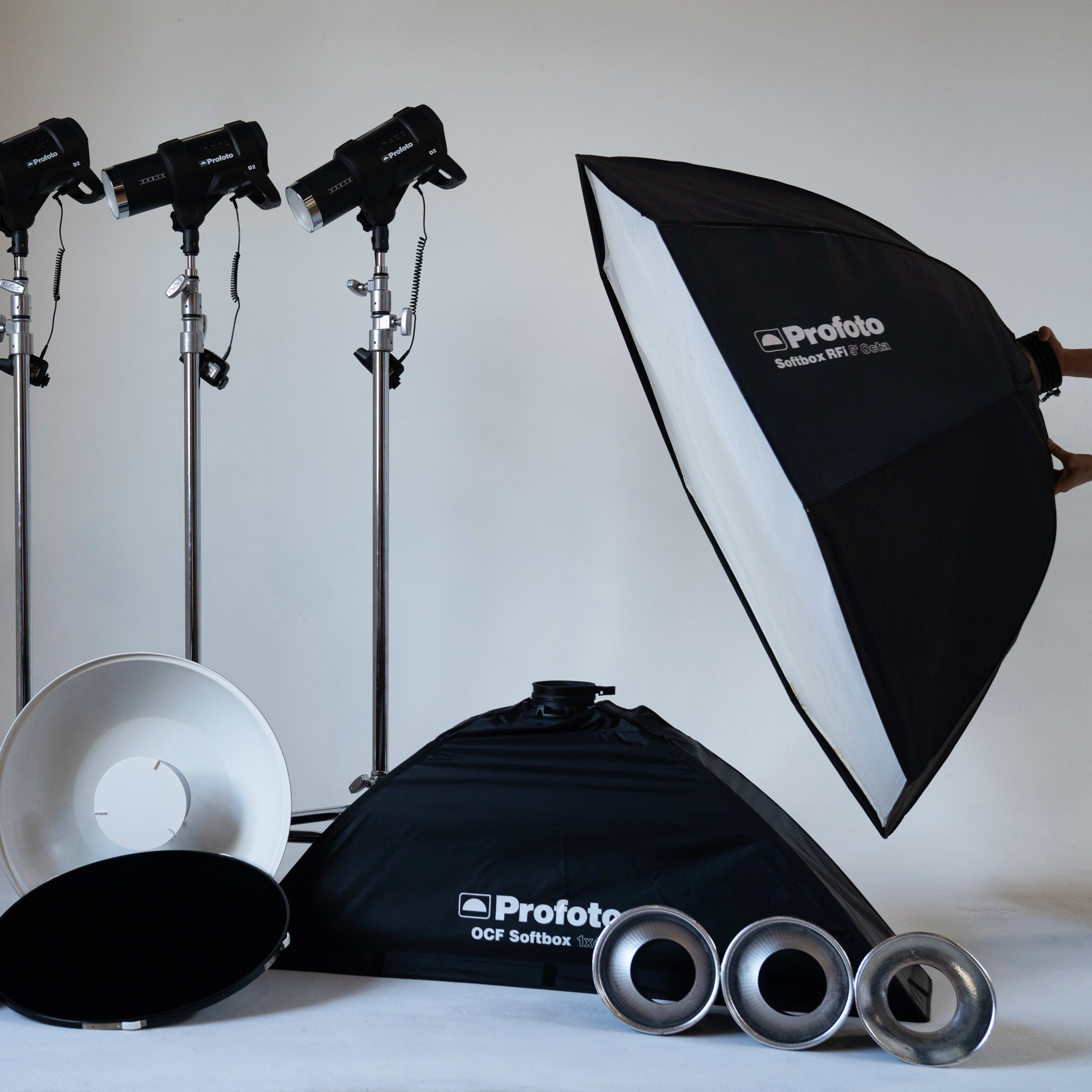 A variety of photography lighting equipment, including three light stands with softboxes, a reflector, and several other lighting accessories arranged against a neutral background.