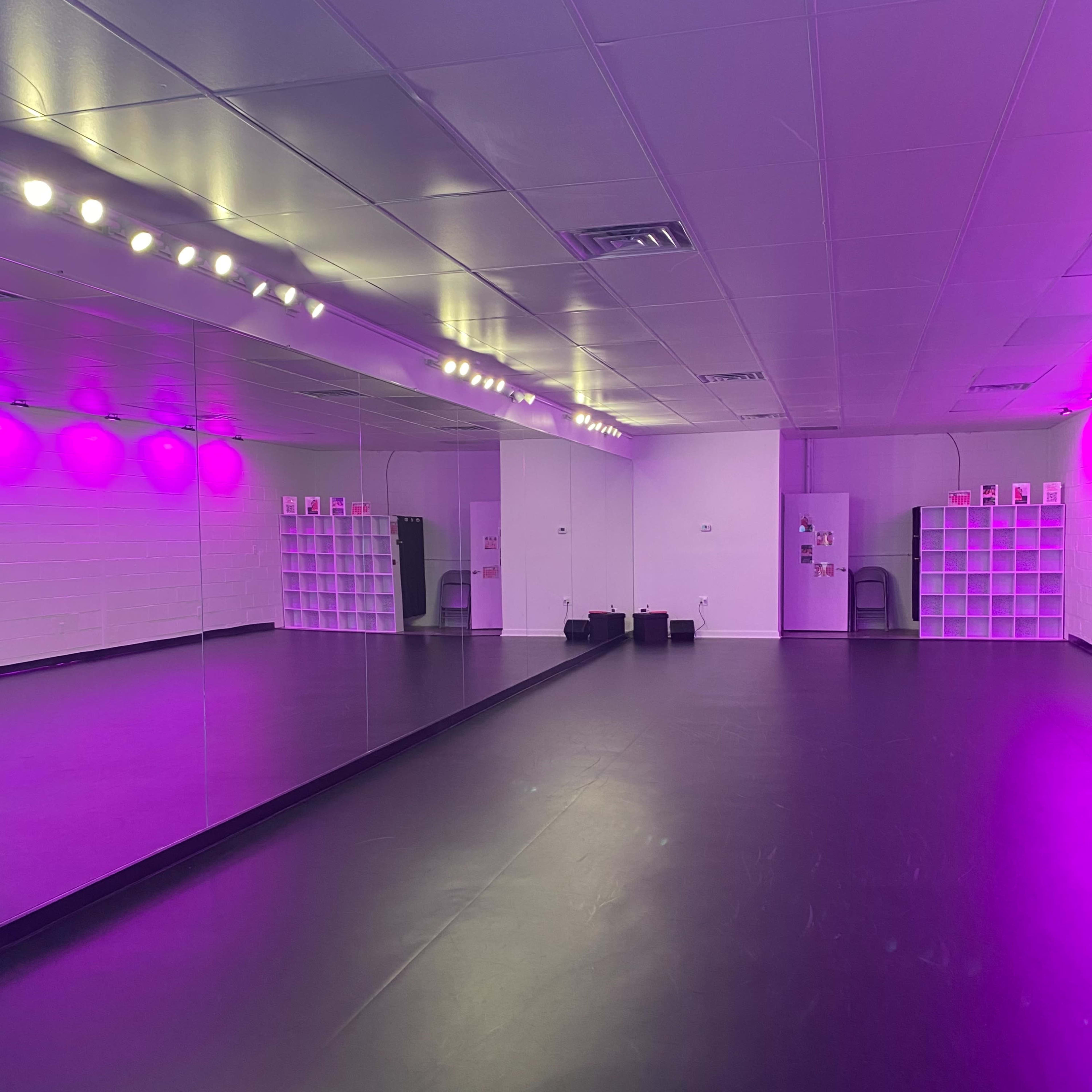 A spacious dance studio with purple lighting, mirrored walls, and black flooring.