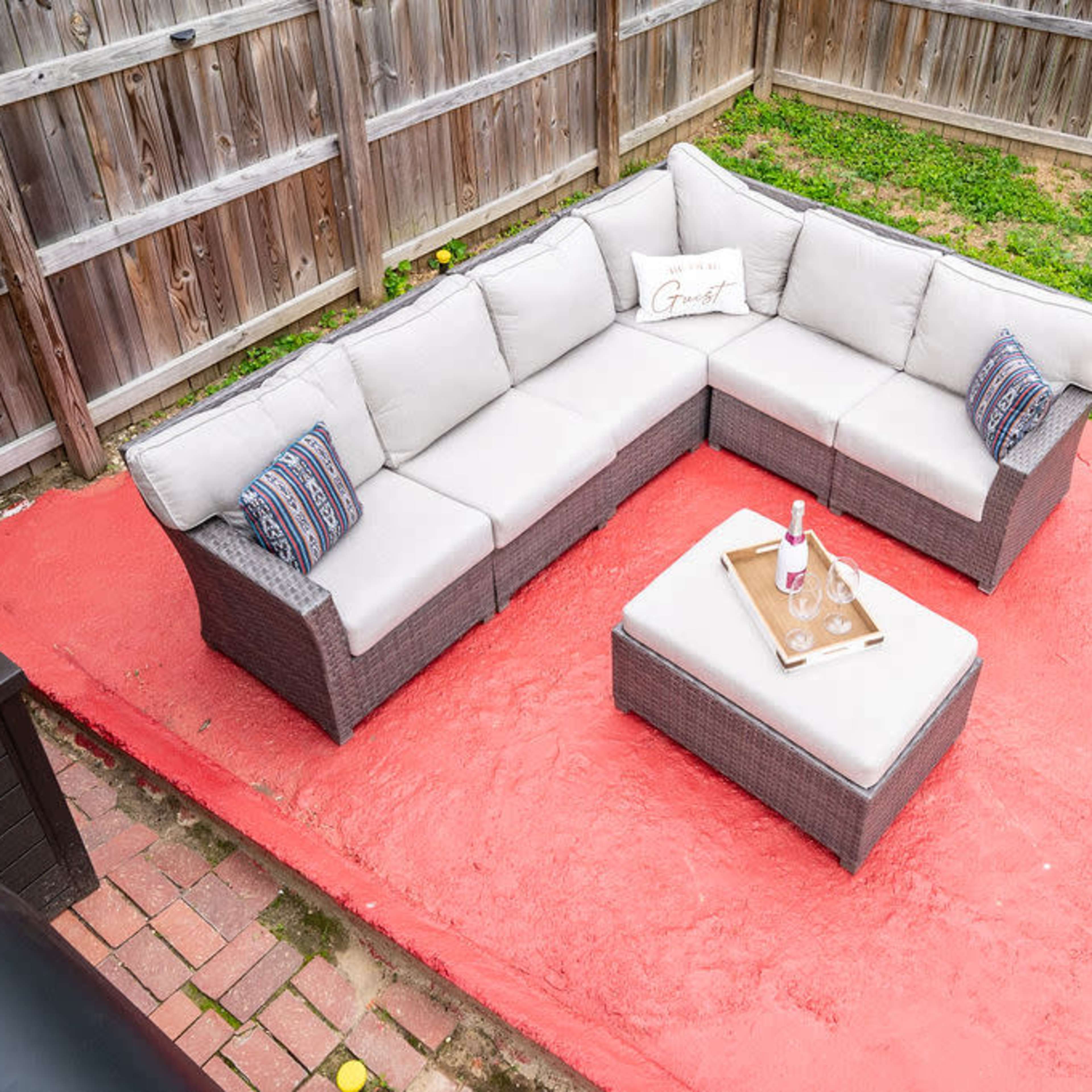A sectional outdoor sofa with a coffee table is placed on a red patio in a fenced backyard.
