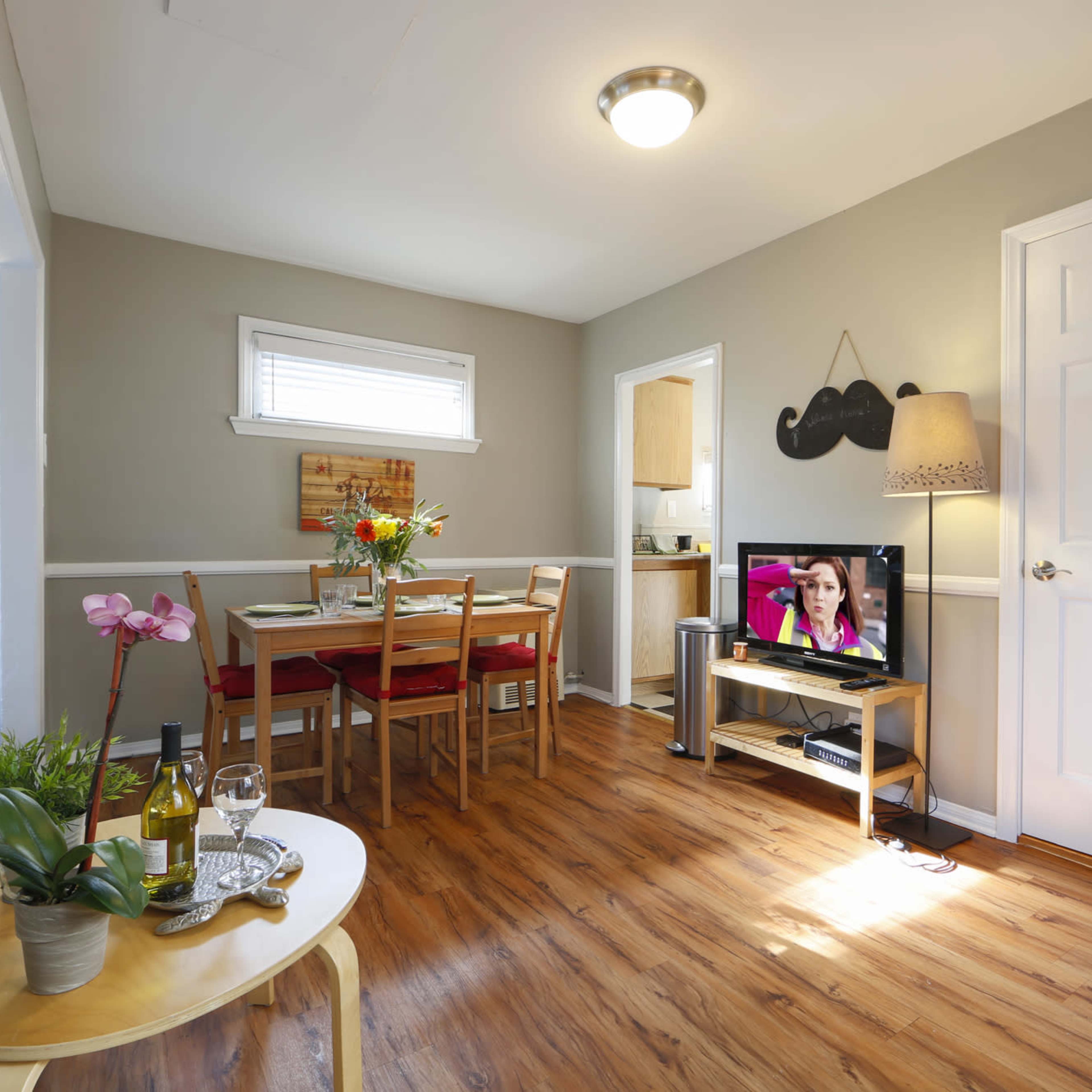A cozy living space features a dining area with a table, chairs, and a television displaying a program, surrounded by light-colored walls and wooden flooring.