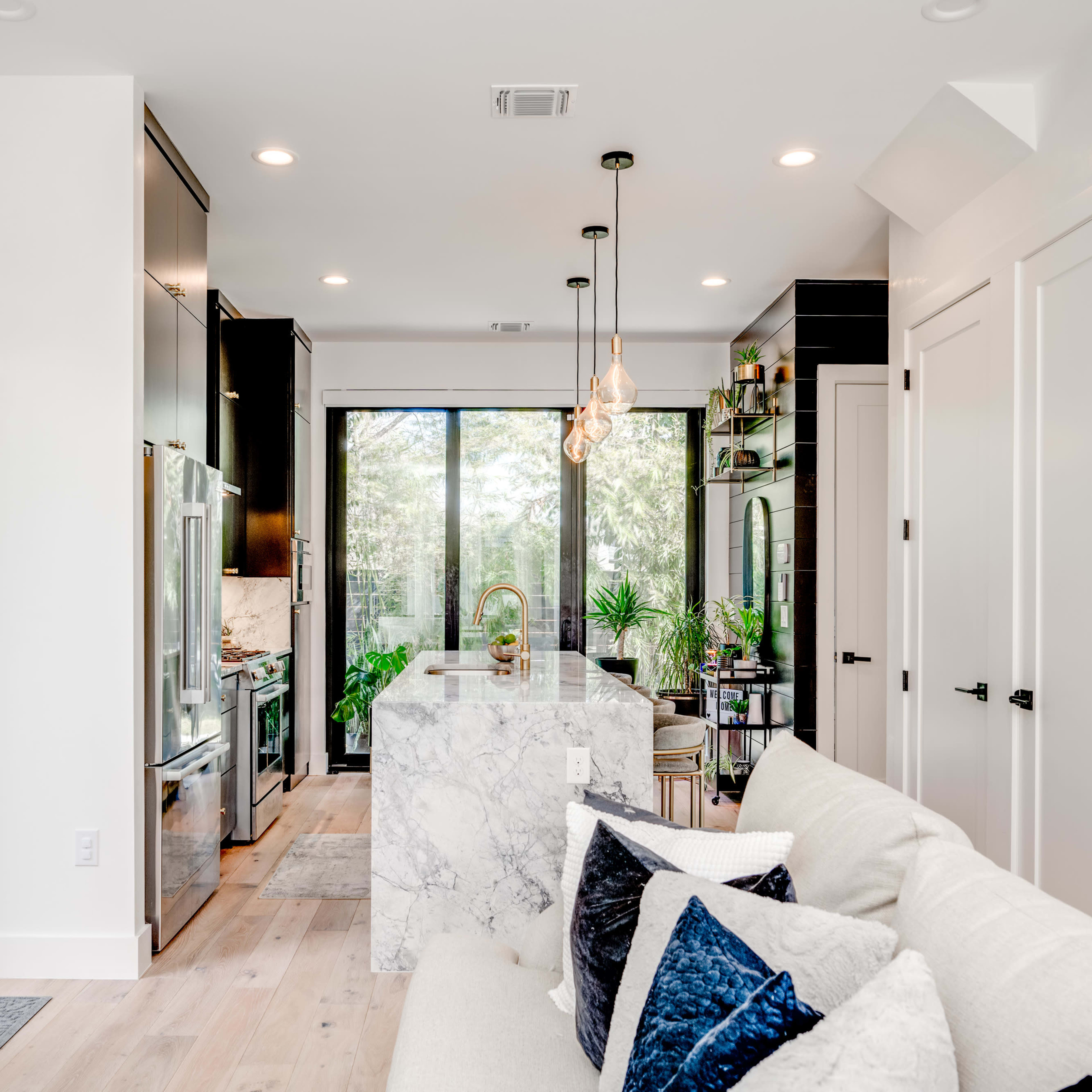 A modern kitchen and living area feature a marble island, sleek cabinetry, and a cozy couch, with large windows allowing natural light and views of greenery.