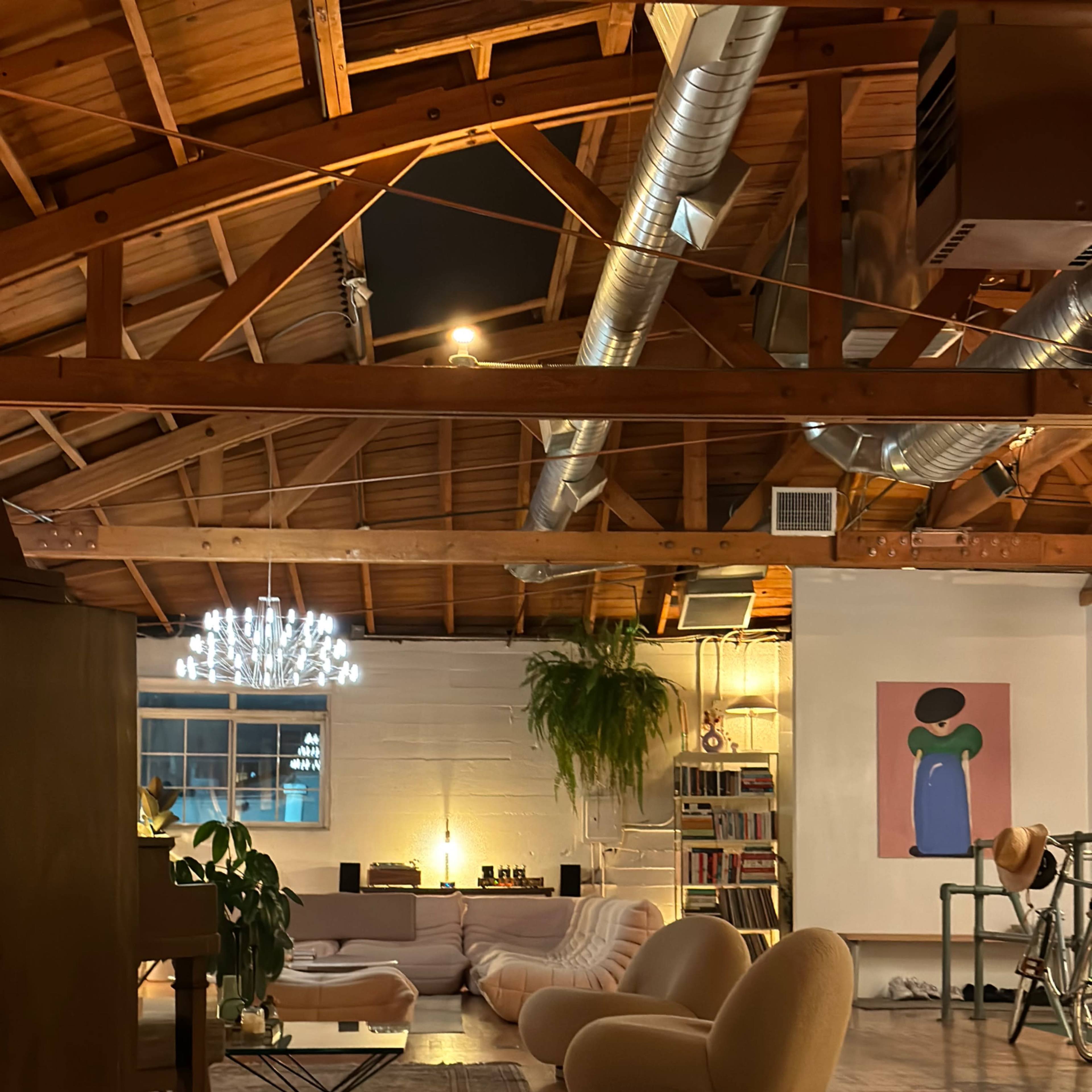 A spacious loft with wooden beams, a large sectional sofa, a round coffee table, and a colorful artwork on the wall, illuminated by soft lighting.